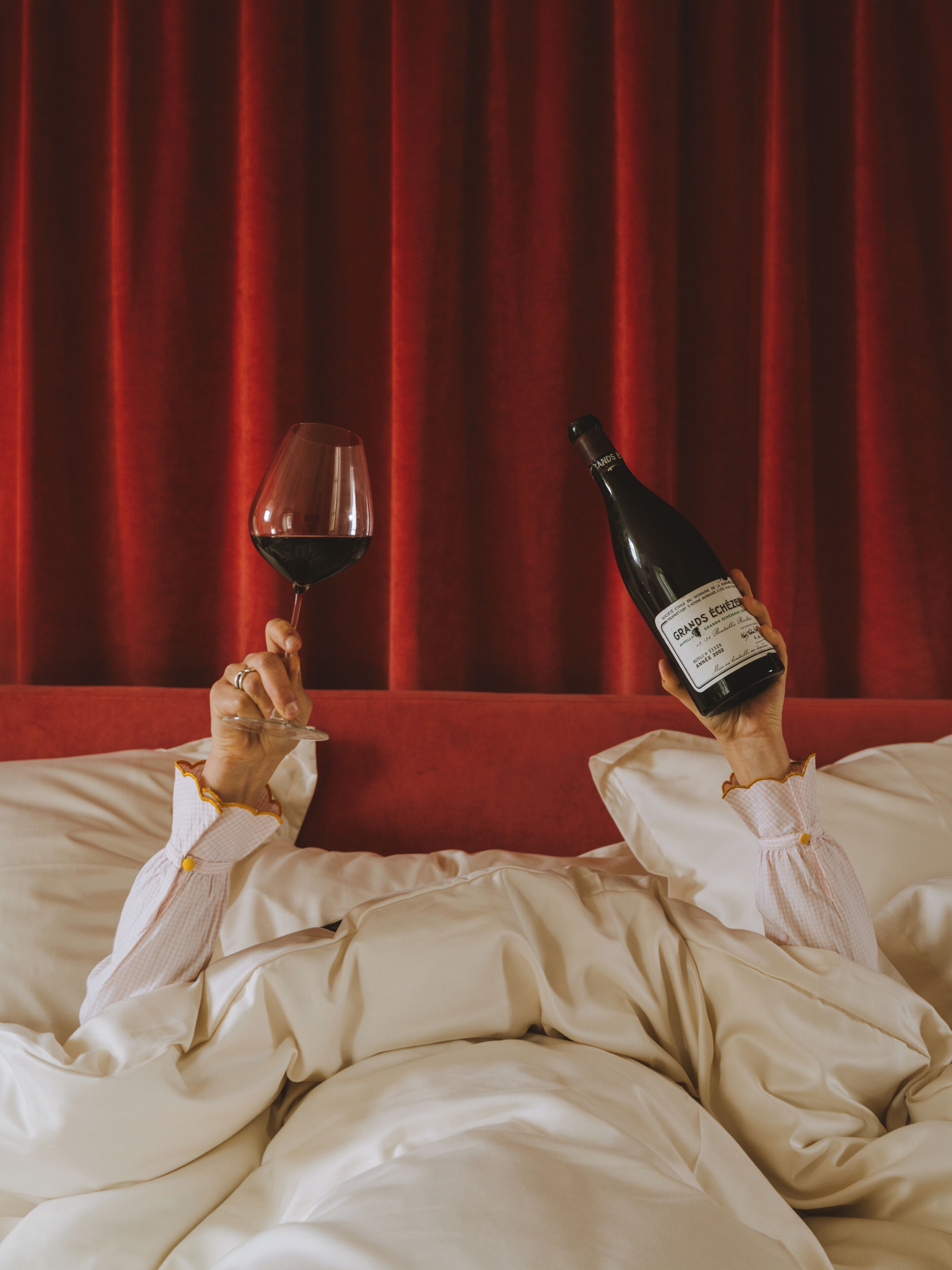 A person reclining on a bed with a bottle of wine and a glass of red wine.