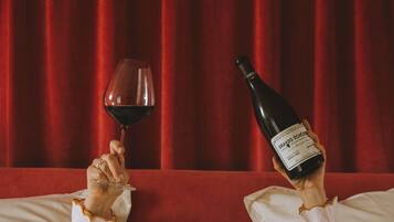 A person reclining on a bed with a bottle of wine and a glass of red wine.