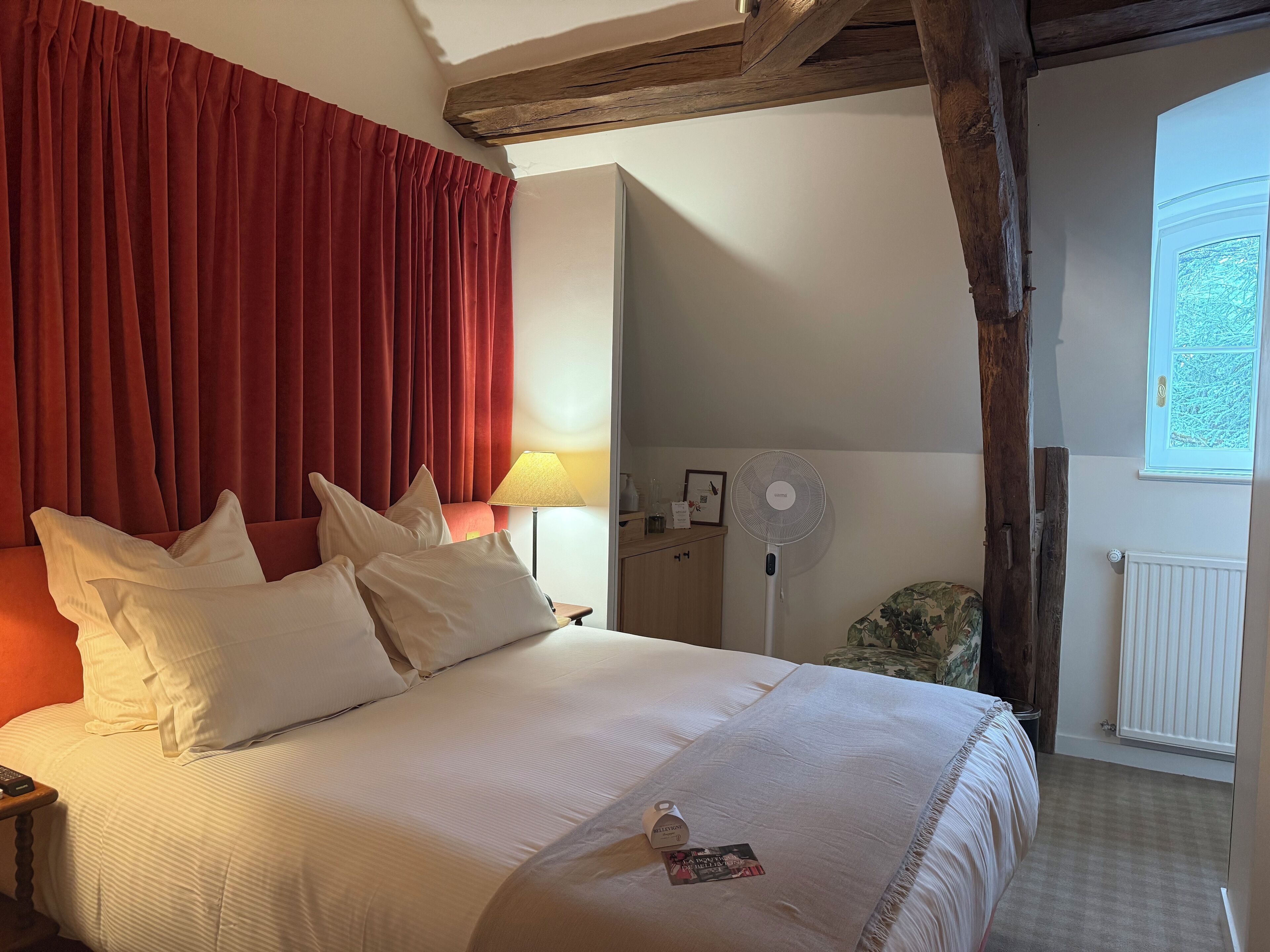 A hotel room with a large bed, red curtains, a bedside lamp, a fan, and a window with a view.