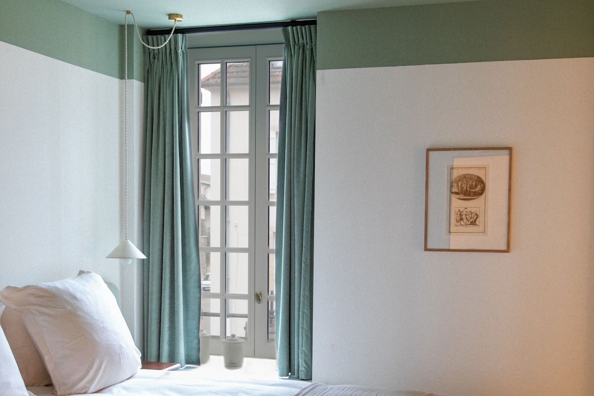 A neatly made bed with a white and peach-colored bedspread, a small round ottoman with a floral pattern, and a framed picture on the wall.