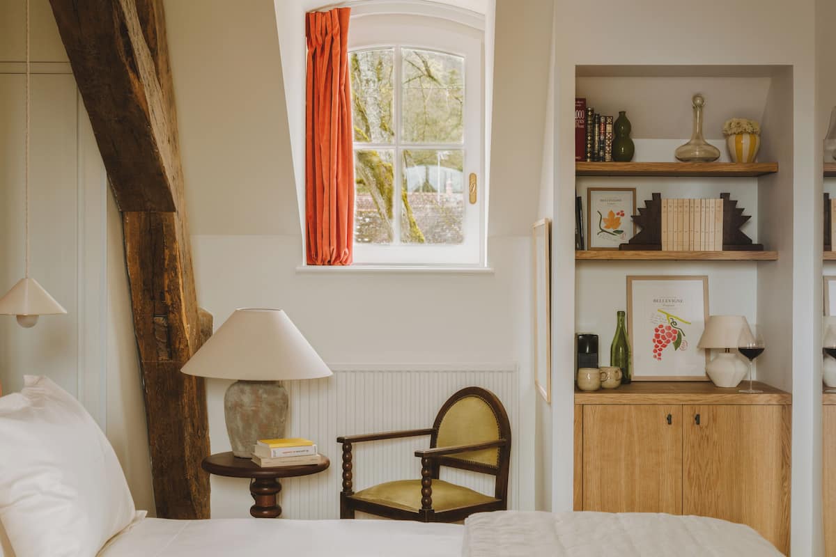 A bedroom with a bed, a chair, a table with books, a window with curtains, and a shelf with books and decorative items.