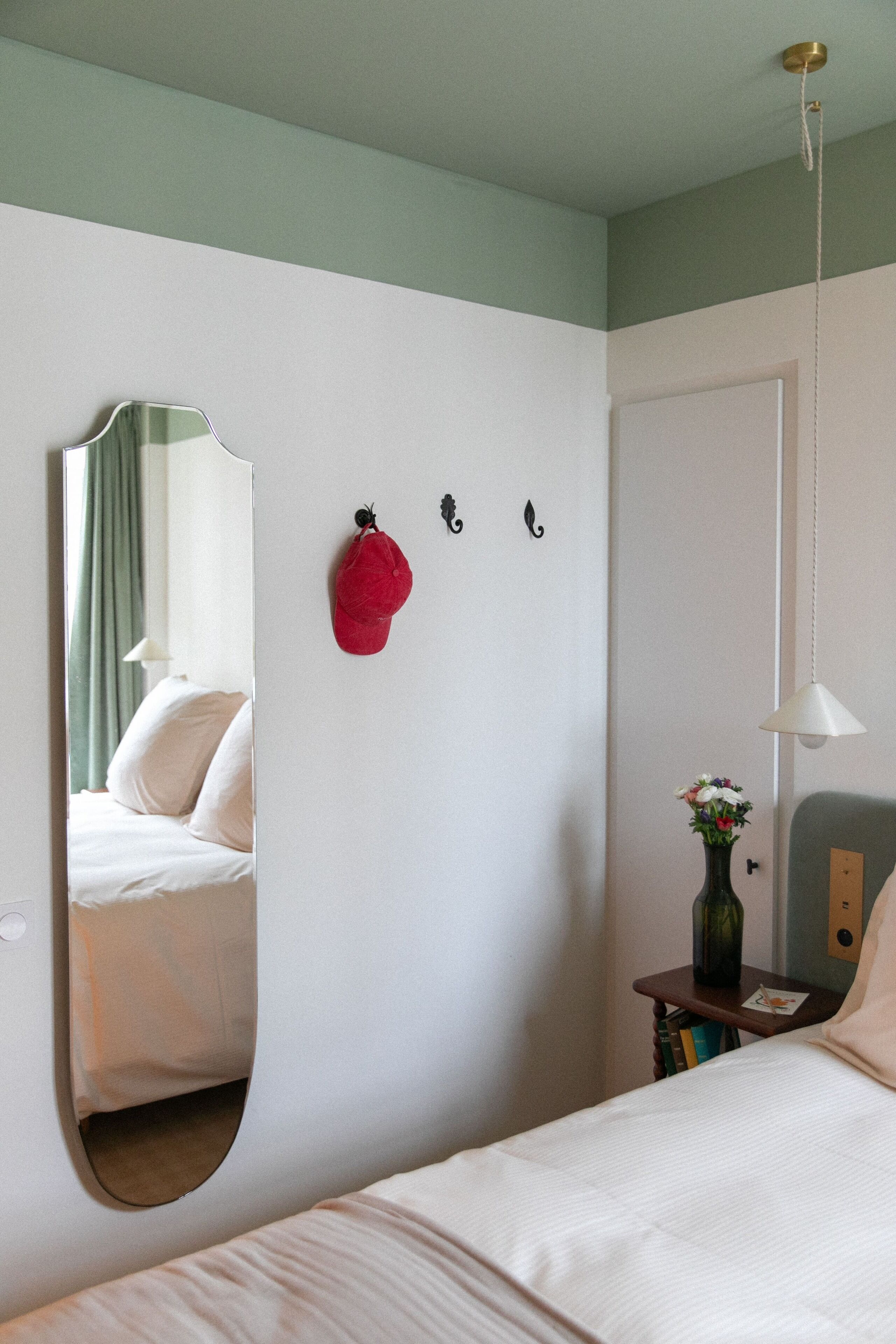 Una habitación de hotel con una cama, un espejo, un sombrero rojo en la pared, una mesita con un jarrón y una lámpara colgante.