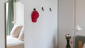 Una habitación de hotel con una cama, un espejo, un sombrero rojo en la pared, una mesita con un jarrón y una lámpara colgante.