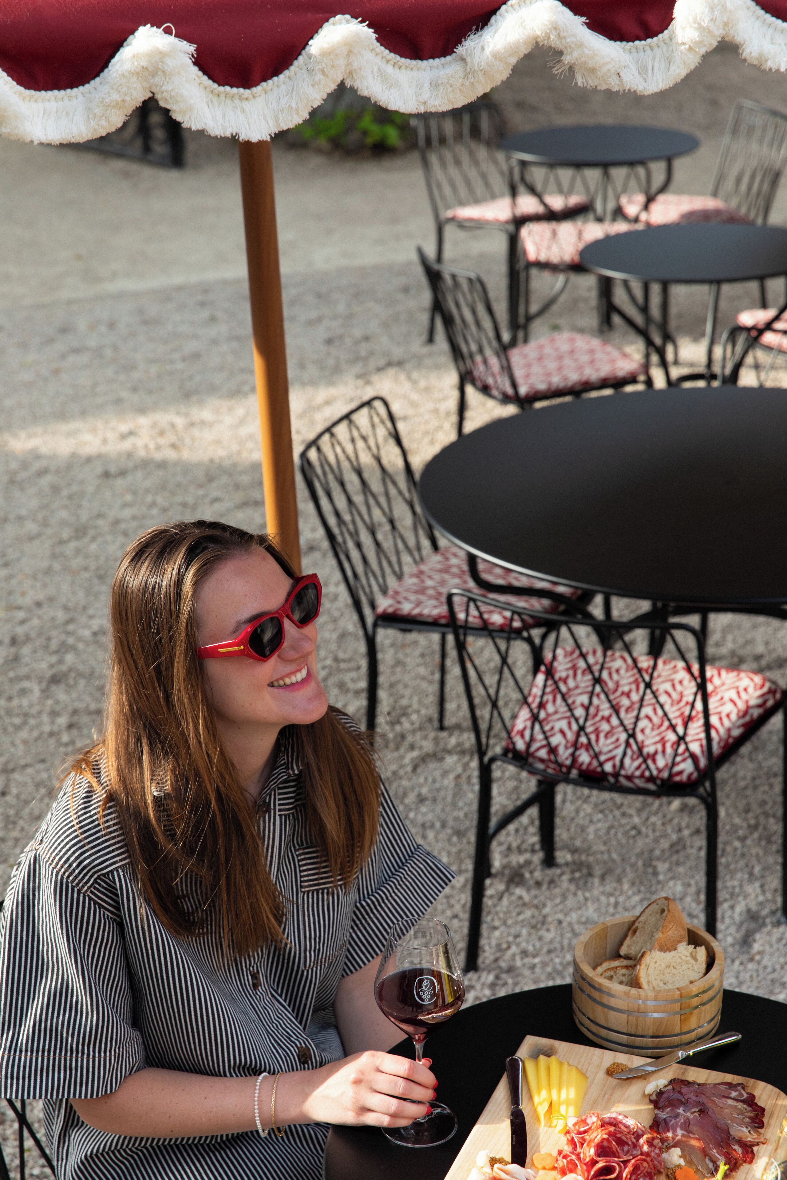 A woman seated at an outdoor table with a wine glass, surrounded by chairs and tables.