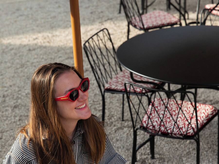 A woman seated at an outdoor table with a wine glass, surrounded by chairs and tables.
