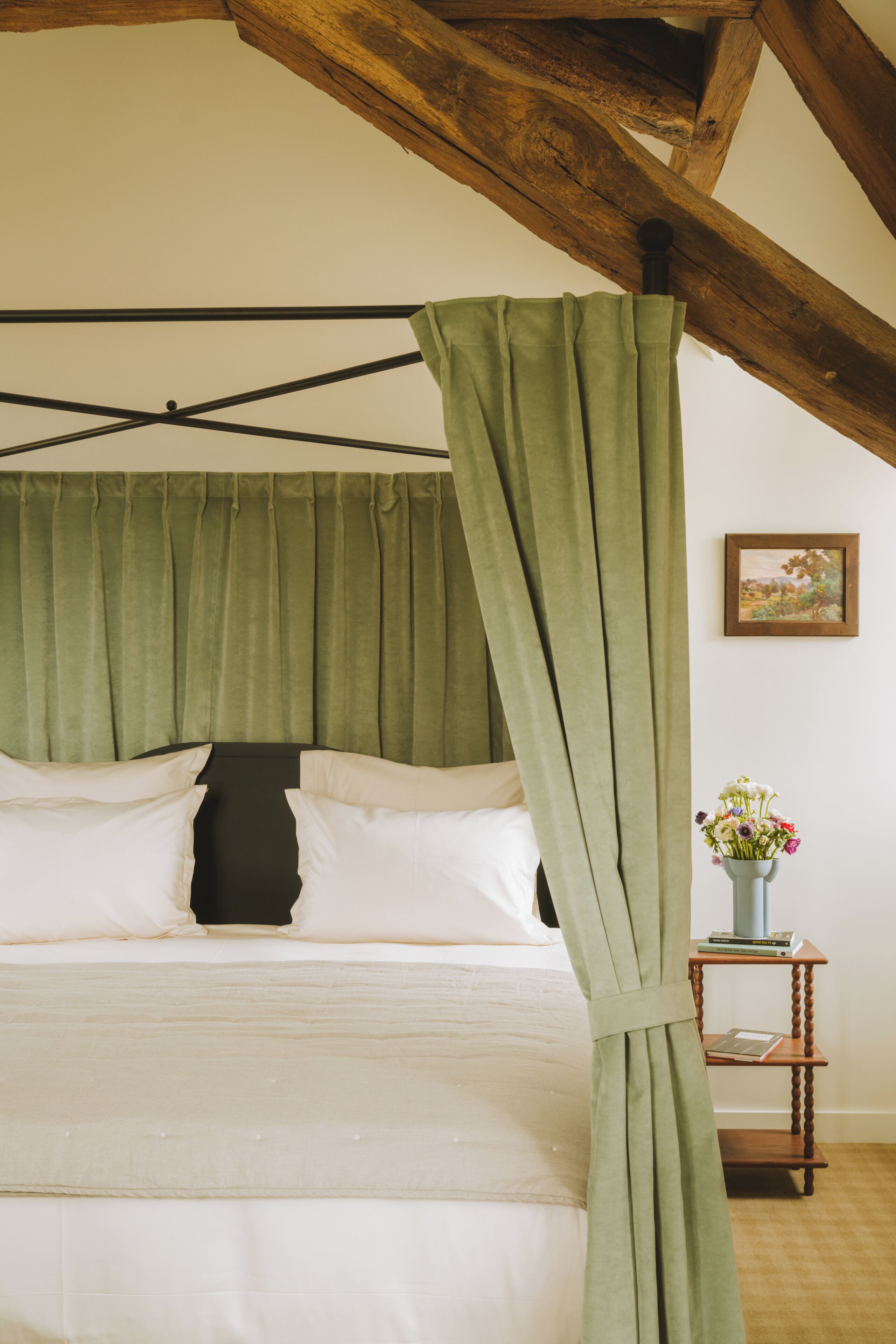 Una cama con cortinas verdes, un cabecero de madera y una mesita con un florero.