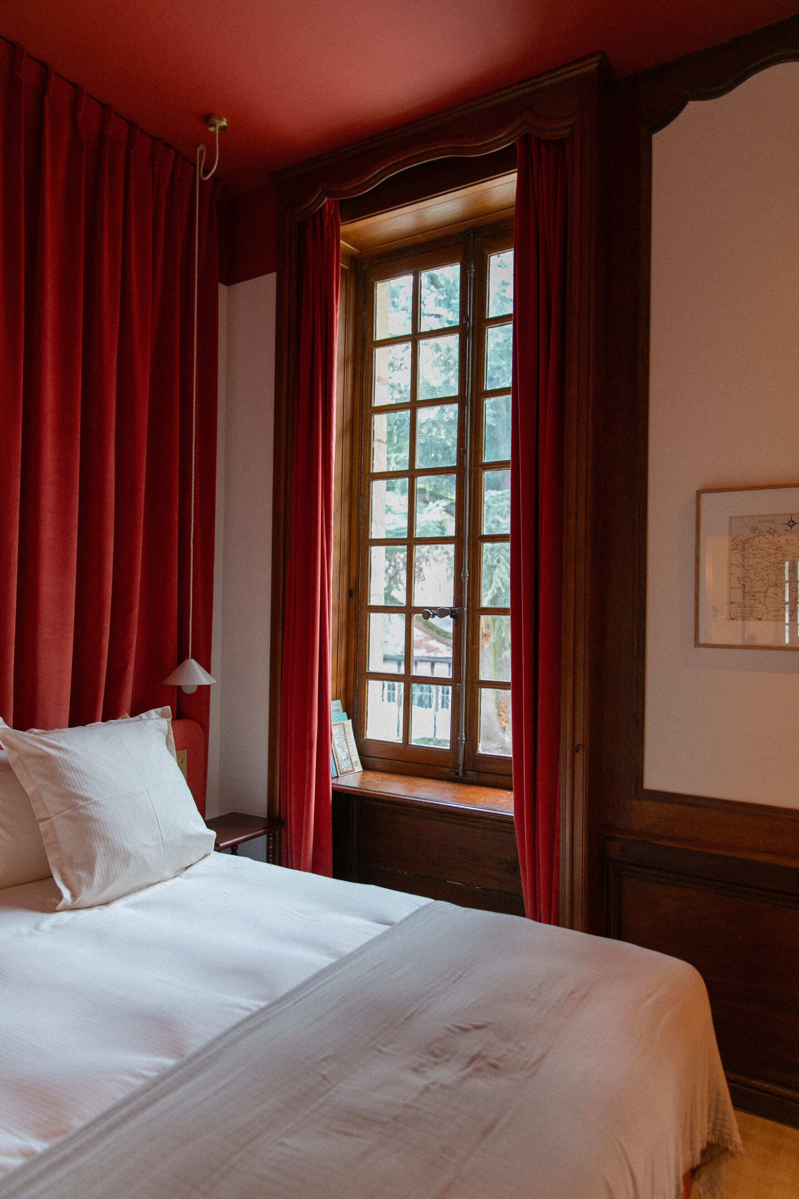 Une chambre d’hôtel avec un grand lit, des rideaux rouges, une fenêtre donnant sur un beau paysage et une carte encadrée accrochée au mur.