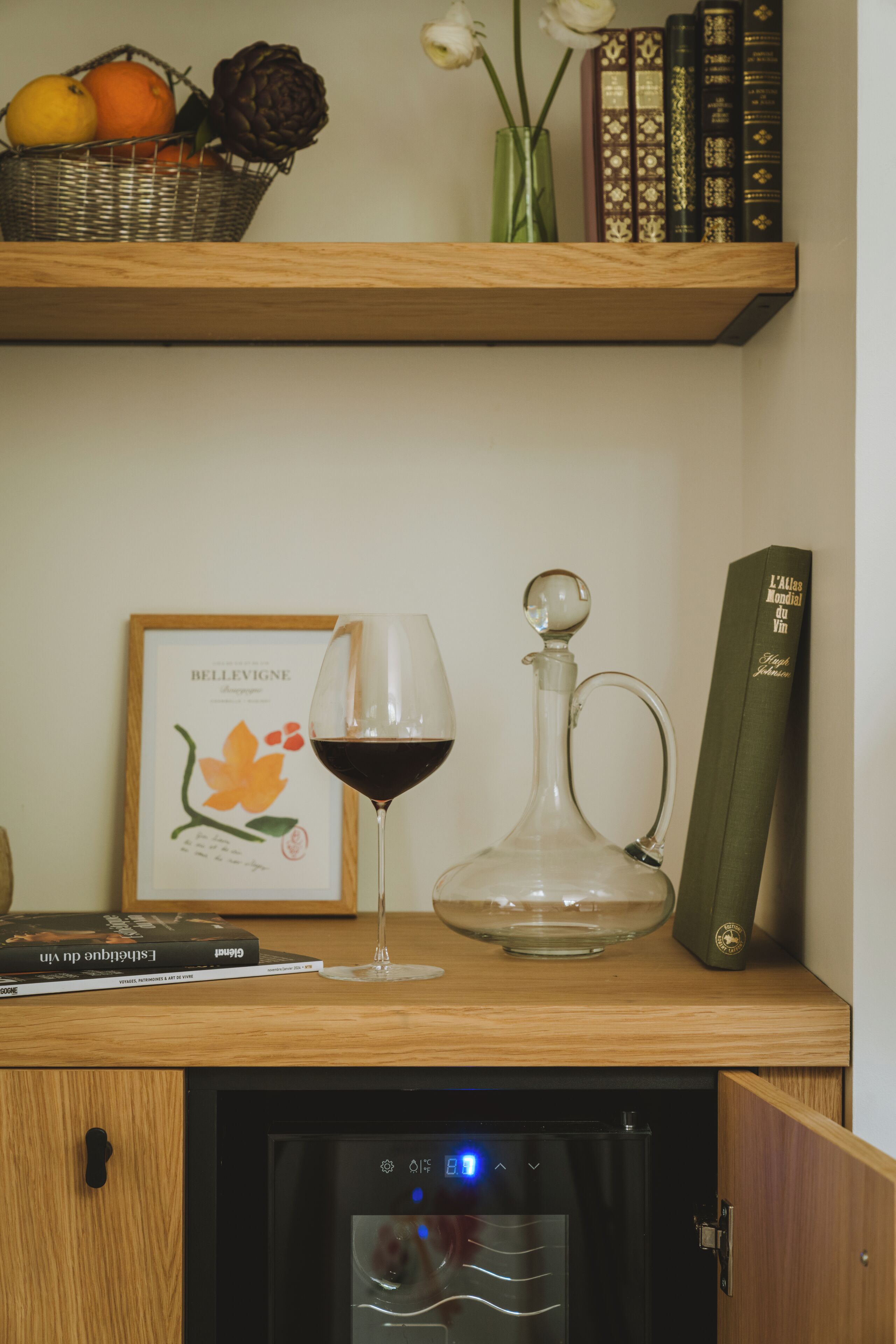 Une étagère en bois sur laquelle on trouve un verre à vin, une carafe, un tableau encadré et des livres.