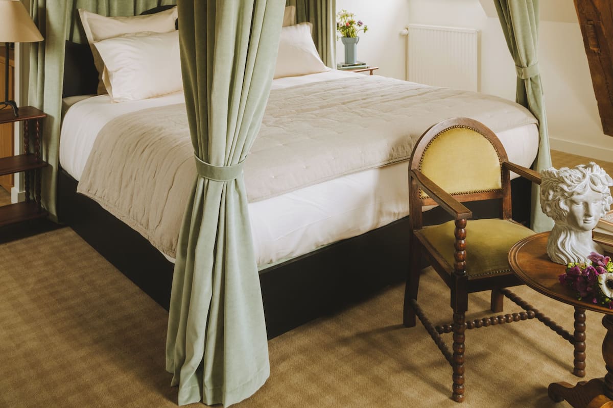 A four-poster canopy bed with green curtains, a wooden chair, and a bust on a small table.
