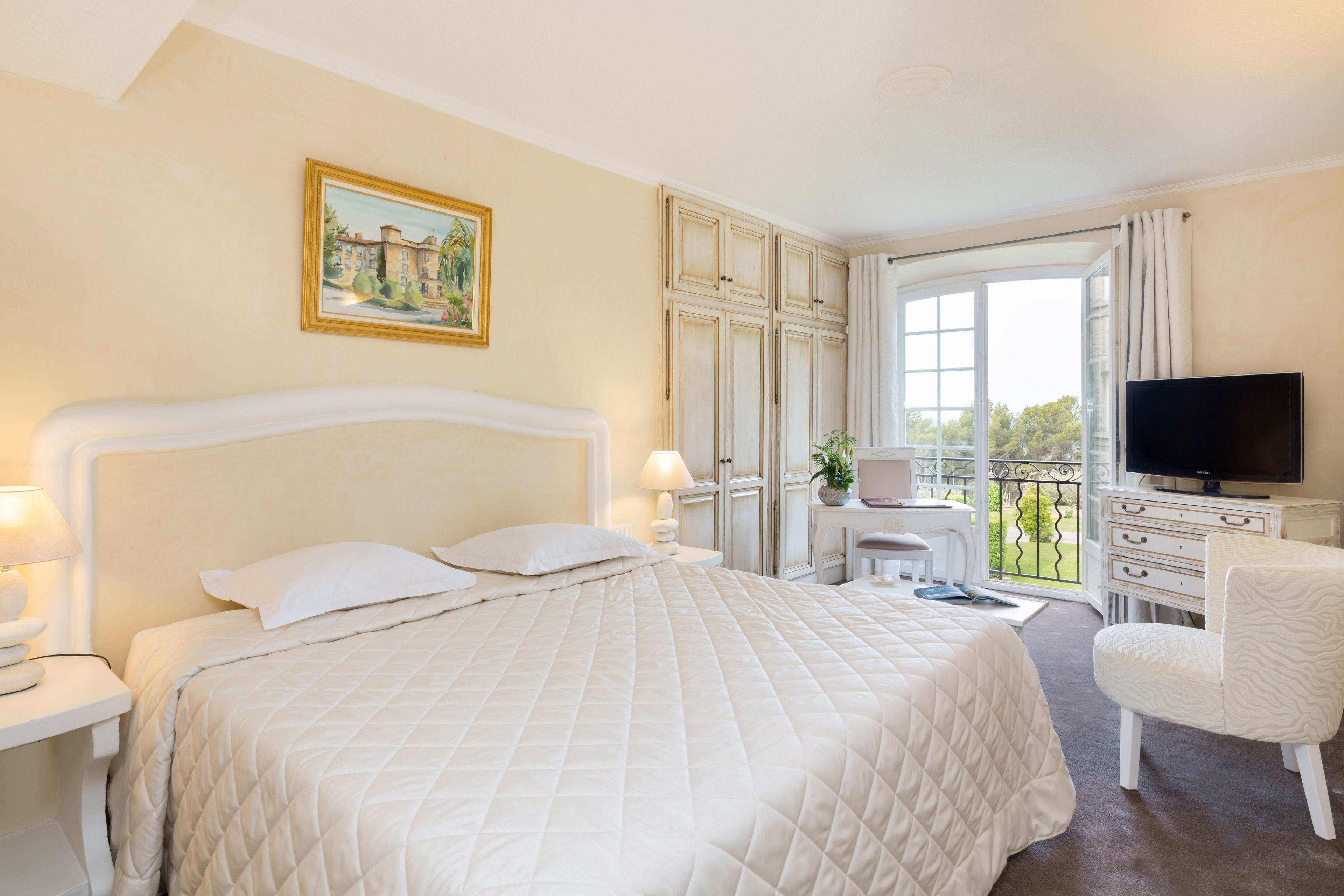 A hotel room with a large bed, a television, a desk with a chair, and a balcony with a view of greenery.
