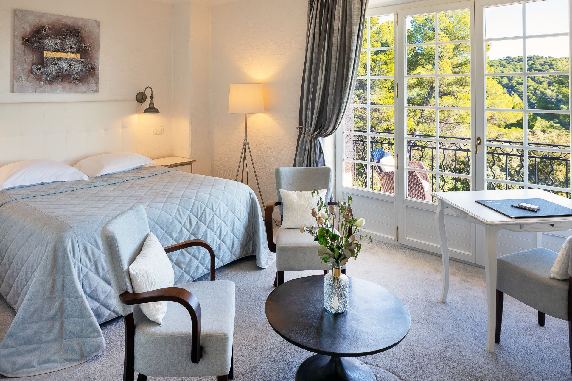 A hotel room with a bed, two chairs, a small table, a lamp, and a window with a view of trees.