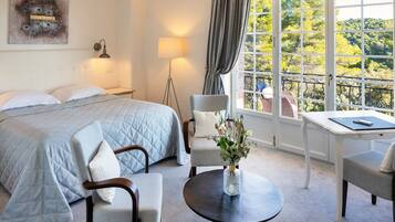 A hotel room with a bed, two chairs, a small table, a lamp, and a window with a view of trees.