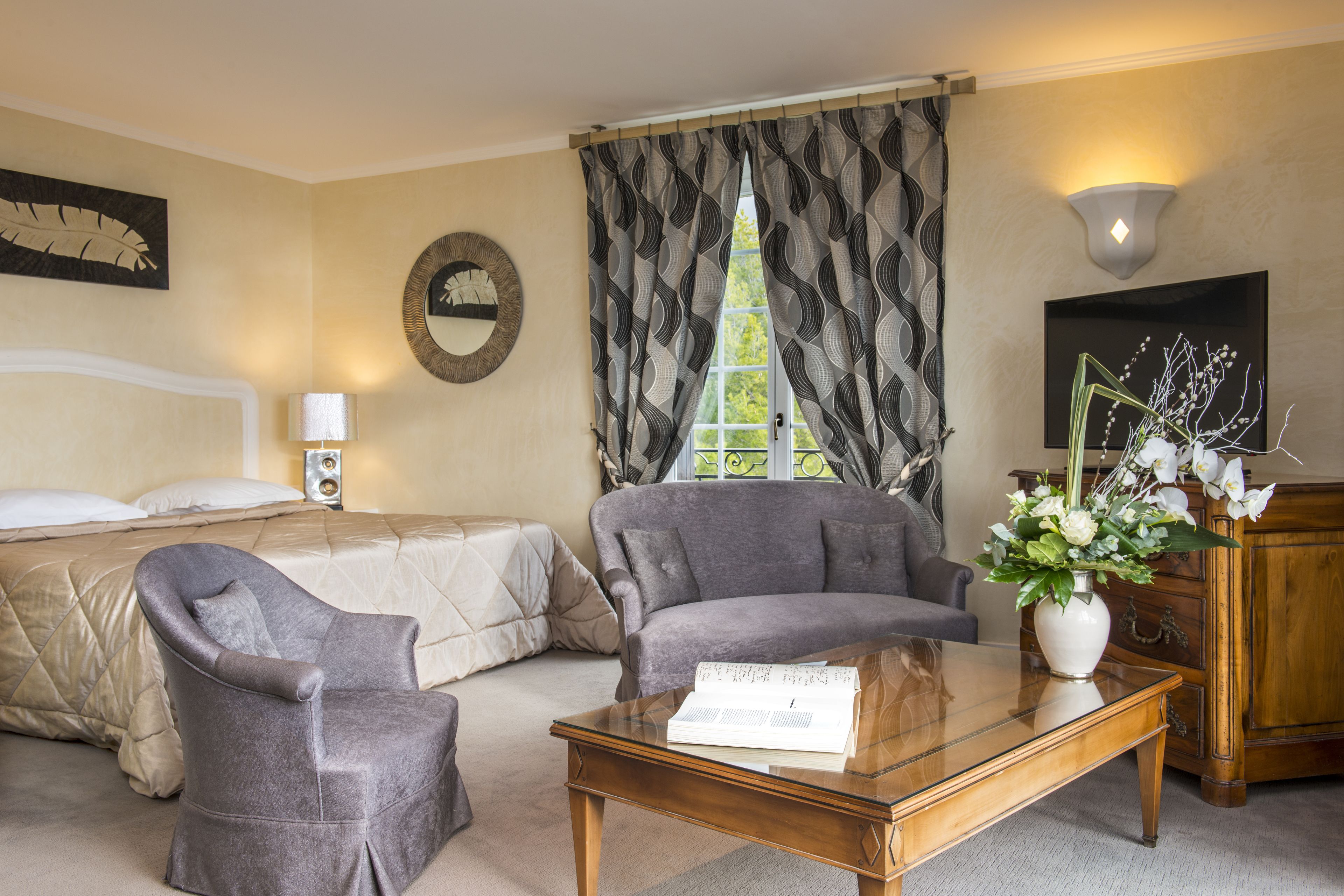 Habitación de hotel con cama, dos sillones, una mesa de centro con un libro, un jarrón con flores, televisión y una ventana con cortinas.