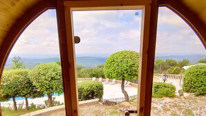 Una terraza de madera con una parrilla Harvia, un banco de madera y vistas a un espacio verde con árboles y un paisaje distante.