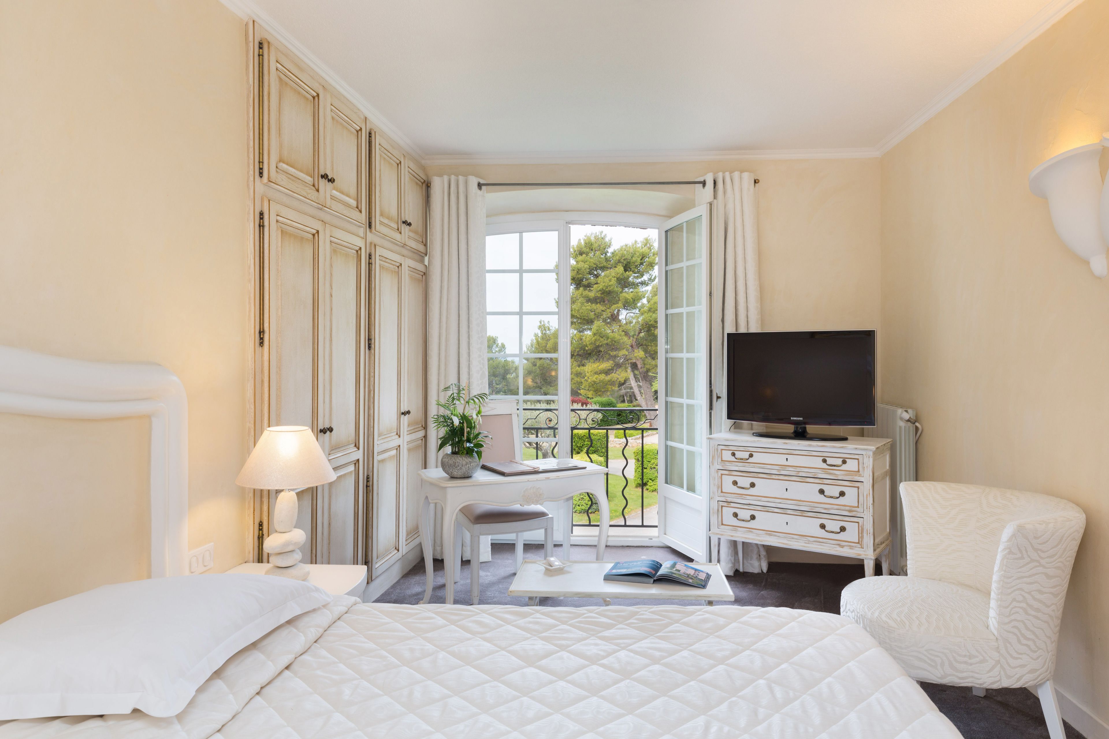 A hotel room with a bed, a desk with a chair, a television, a dresser, and a balcony with a view of trees.