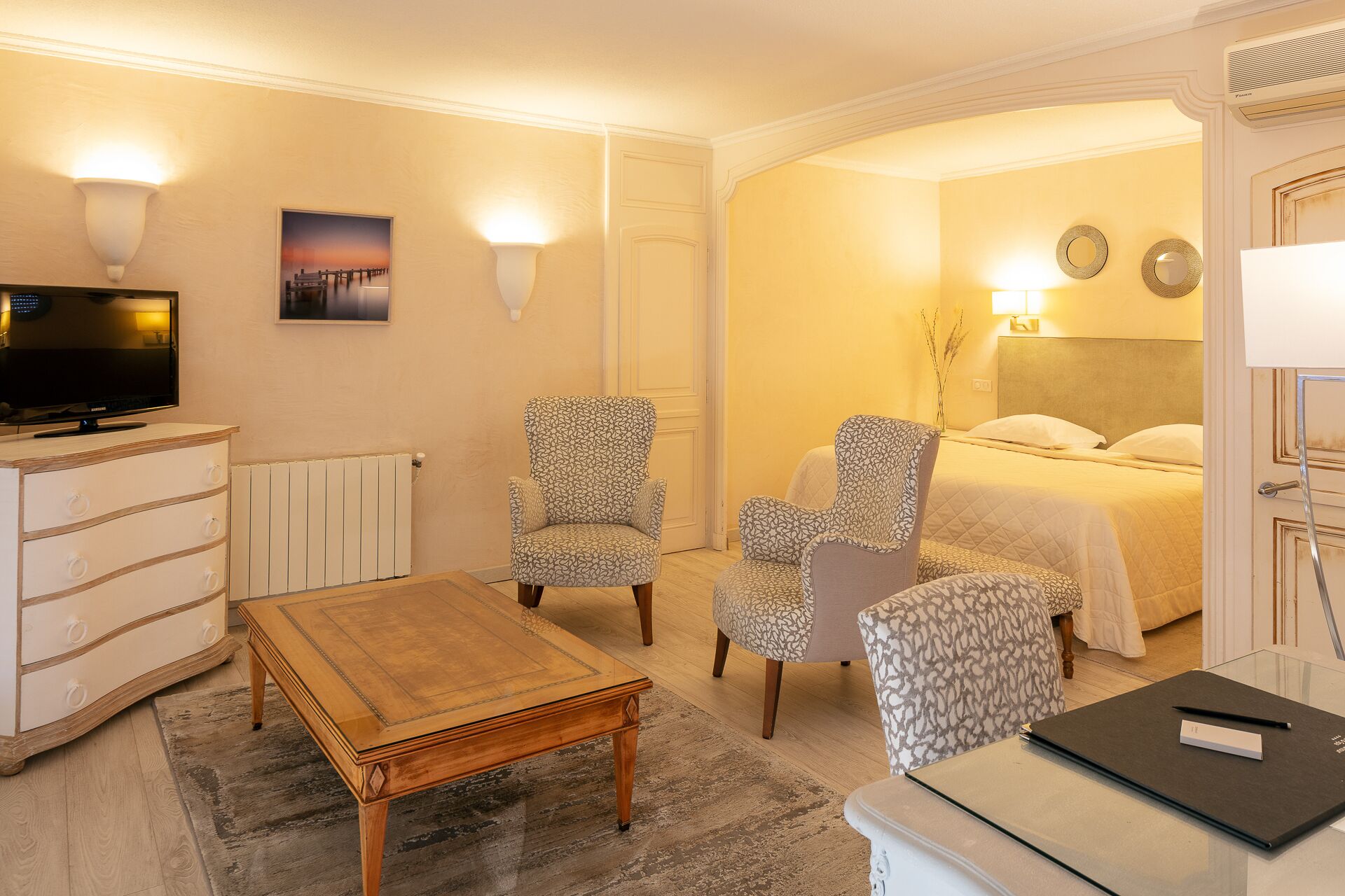 A hotel room with a bed, two armchairs, a wooden coffee table, a TV, and a desk with a laptop.