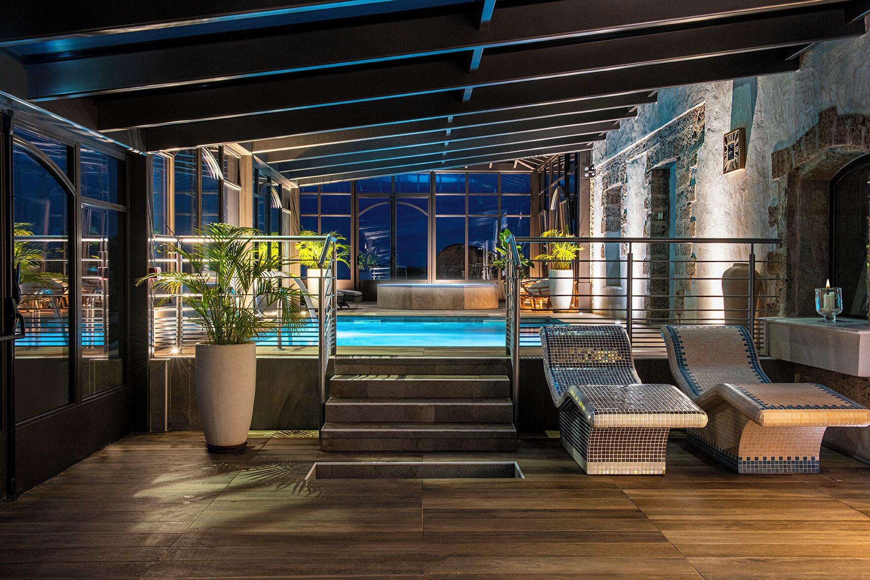 Un área de piscina interior moderna con vistas a la ciudad de noche, que cuenta con una piscina, sillas de descanso y una escalera que conduce a un nivel superior.