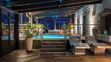 Un área de piscina interior moderna con vistas a la ciudad de noche, que cuenta con una piscina, sillas de descanso y una escalera que conduce a un nivel superior.
