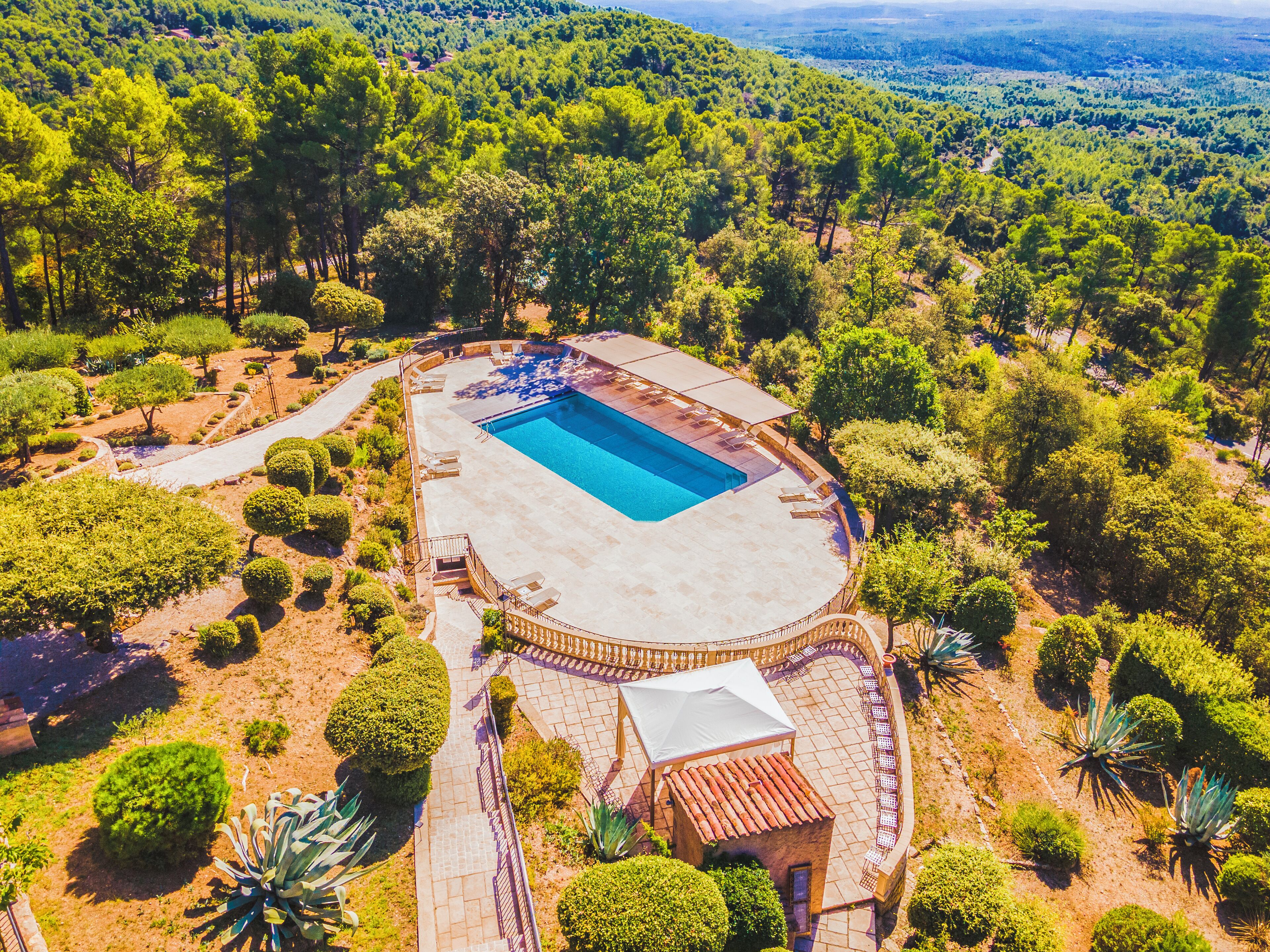 Vue aérienne d’un complexe hôtelier doté d’une piscine, entouré de jardins aménagés, avec en arrière-plan les collines boisées environnantes.