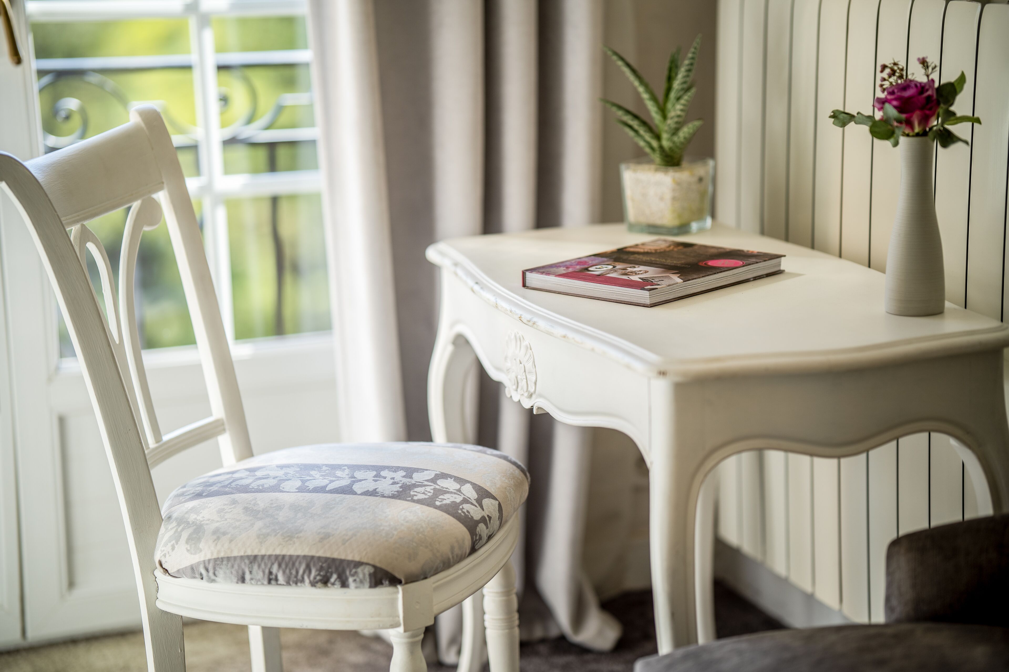 Una sedia bianca con un cuscino floreale, un tavolo bianco con un libro e un vaso con fiori, e una finestra con tende.