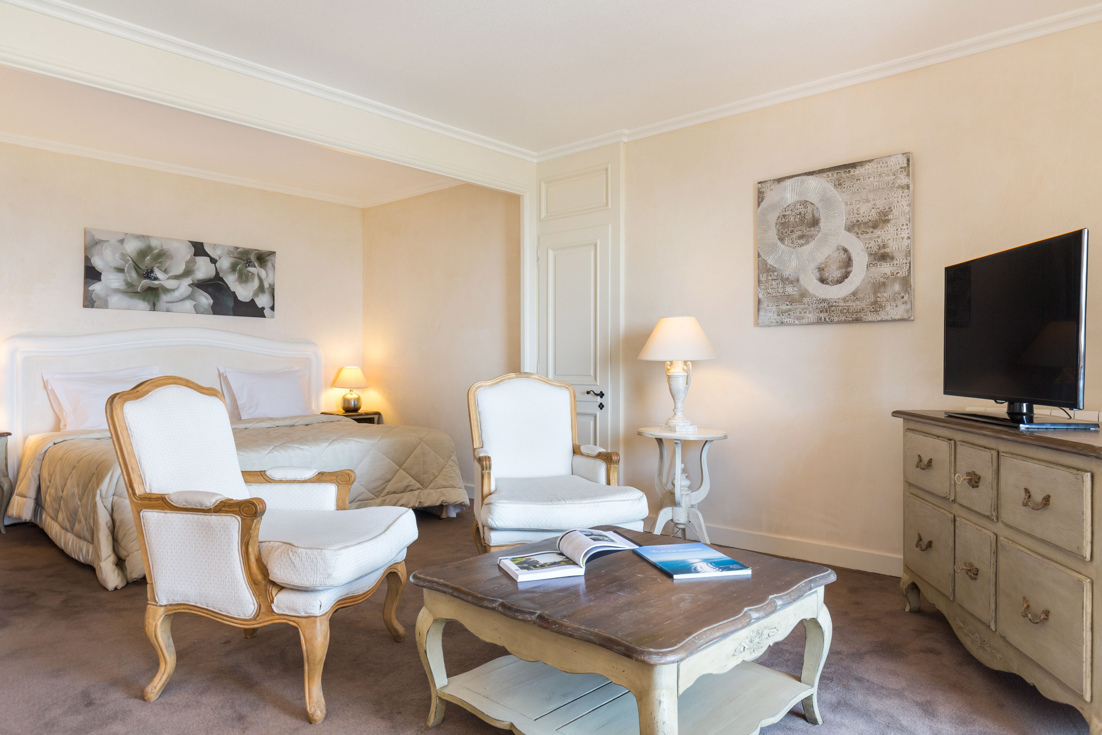 A hotel room with a bed, two white armchairs, a small table with a book, a lamp, a television, and artwork on the walls.
