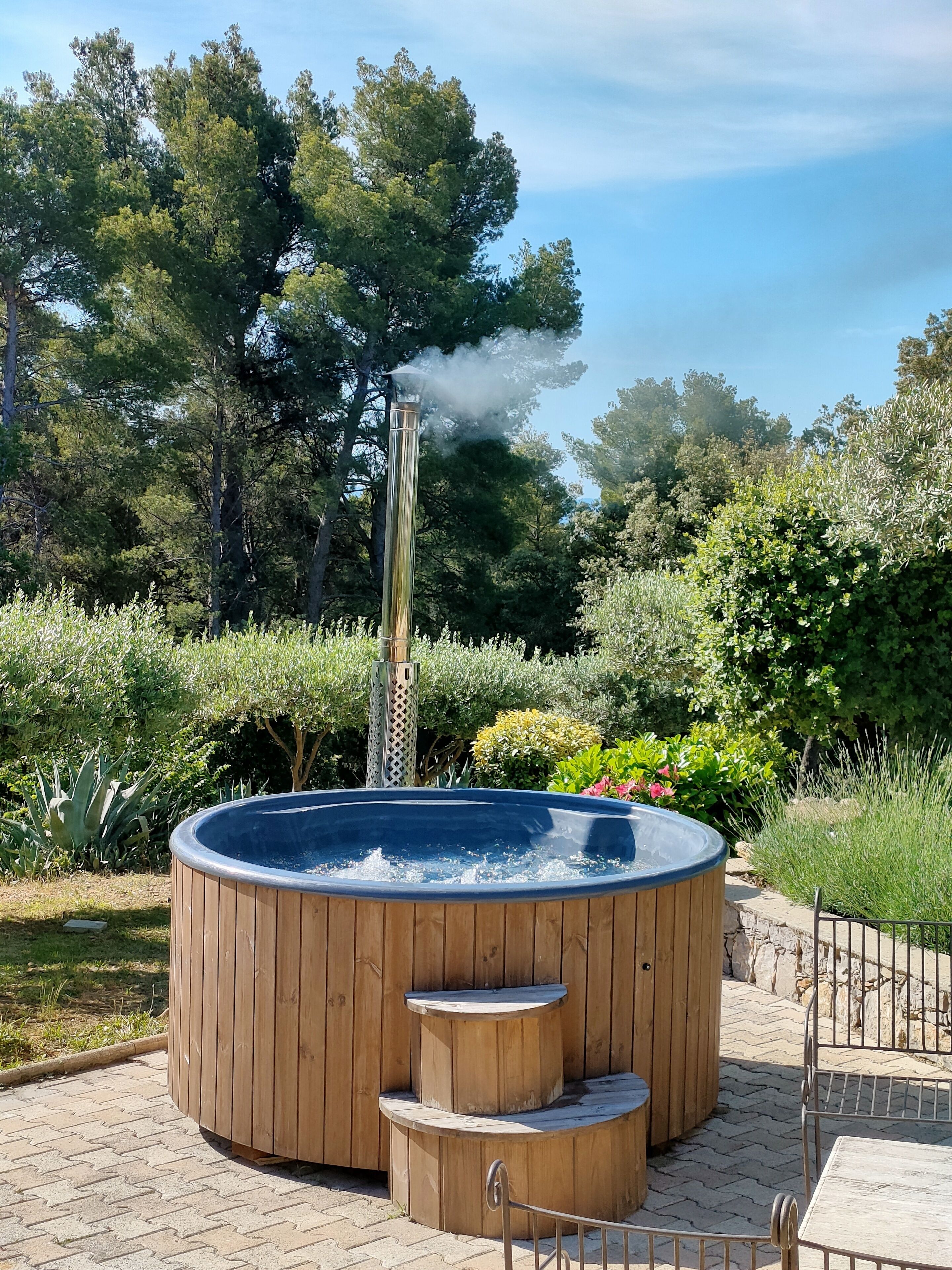 A hot tub with a wooden exterior and a chimney emitting smoke, surrounded by lush greenery and a paved area.