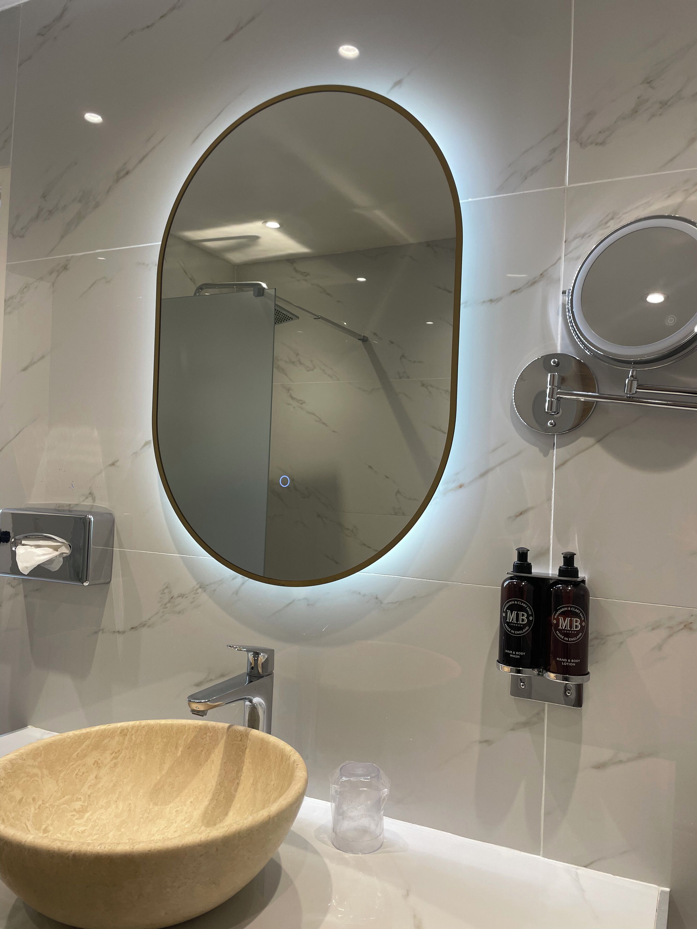 A bathroom with a marble-tiled wall, a round mirror with a light, a wooden bowl sink, and two bottles of hand and body wash.