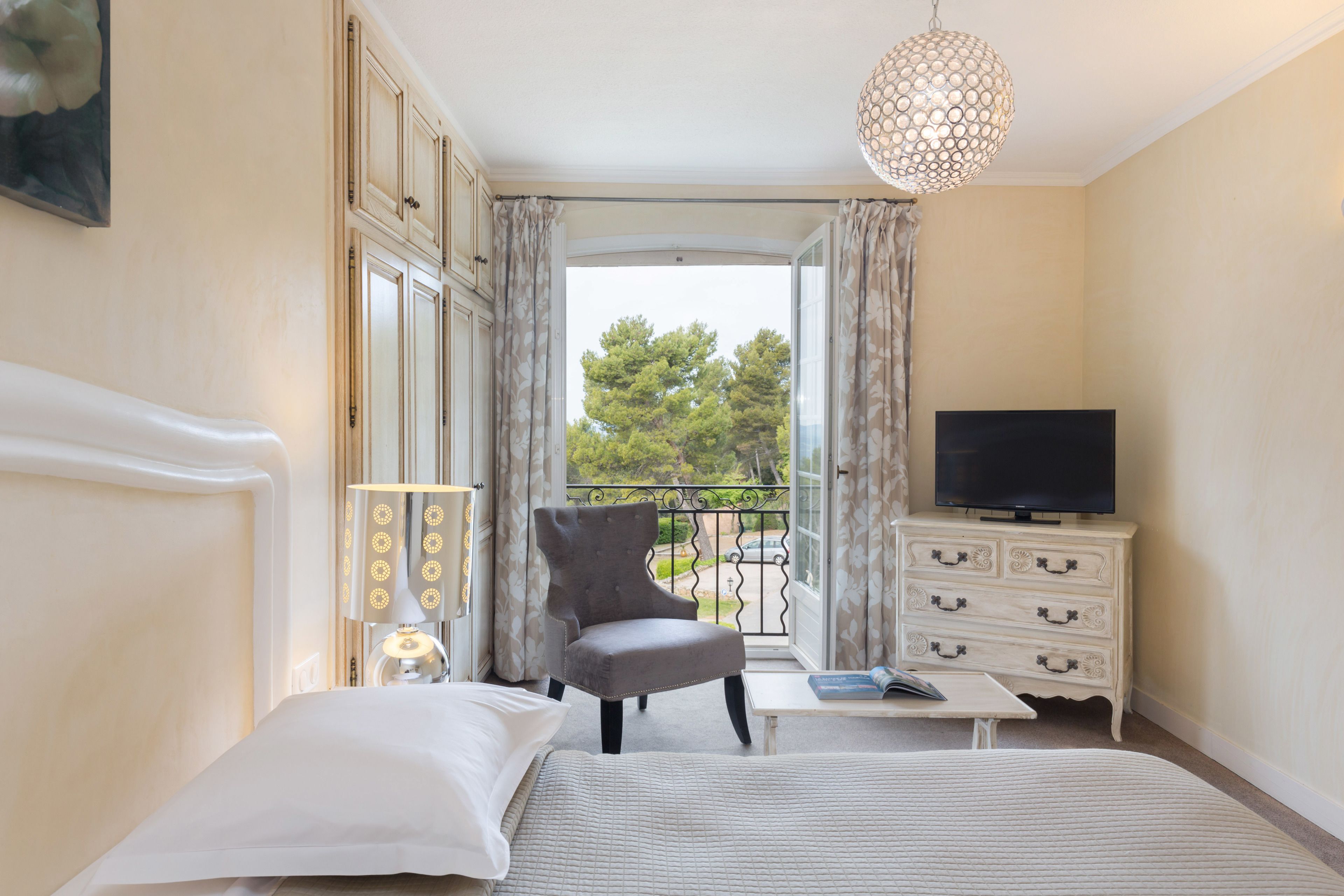A hotel room with a bed, a chair, a TV, a dresser, and a balcony with a view of trees.