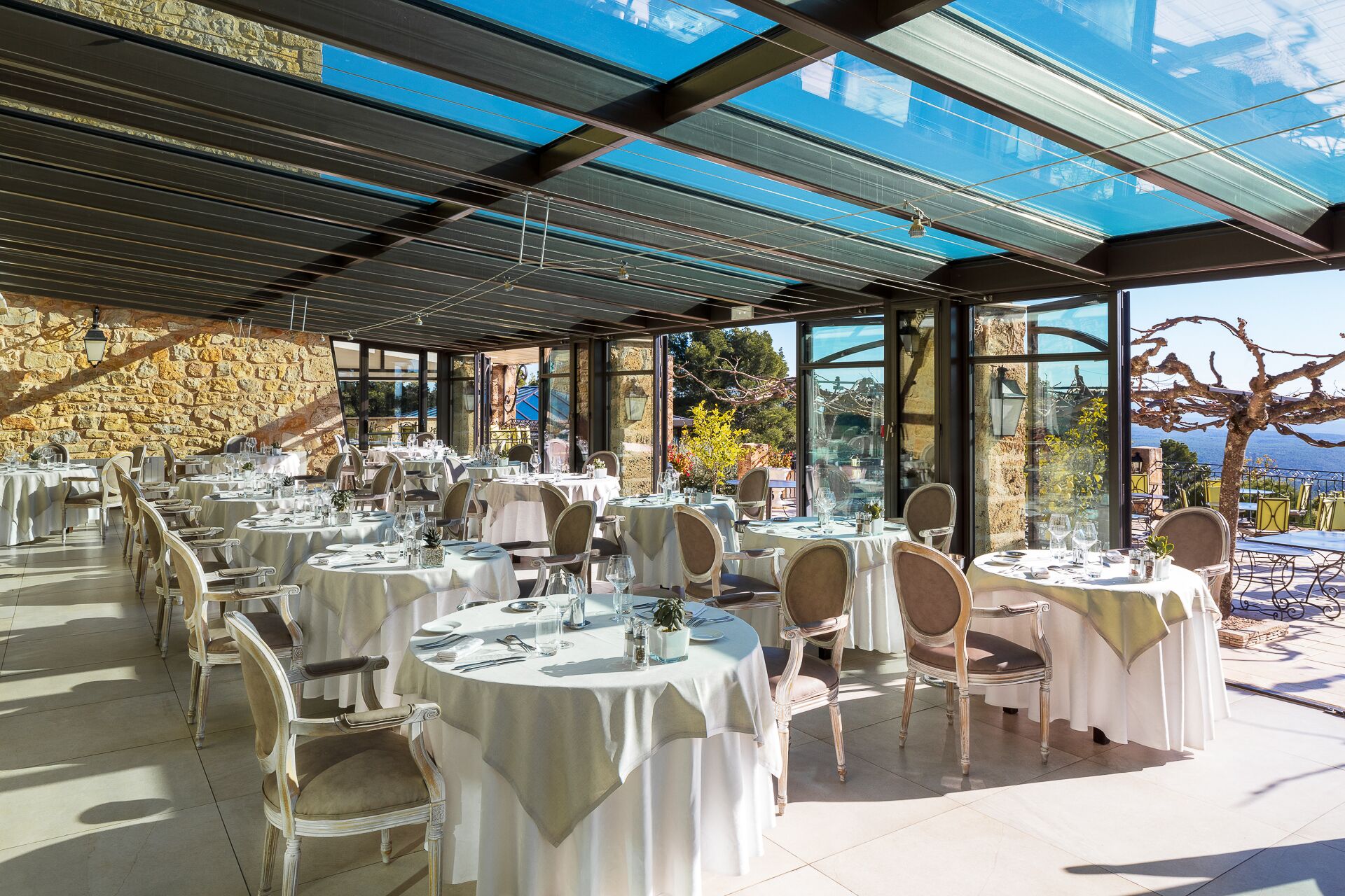Een restaurant met ronde tafels en stoelen, grote ramen en een stenen muur.