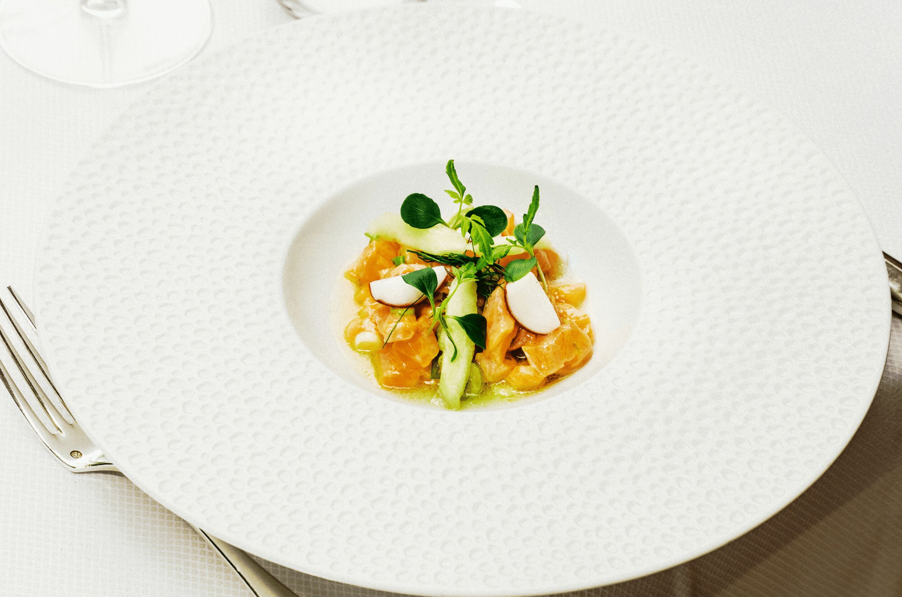 Une assiette blanche contenant un plat, une fourchette et un verre d’eau, le tout posé sur une table.