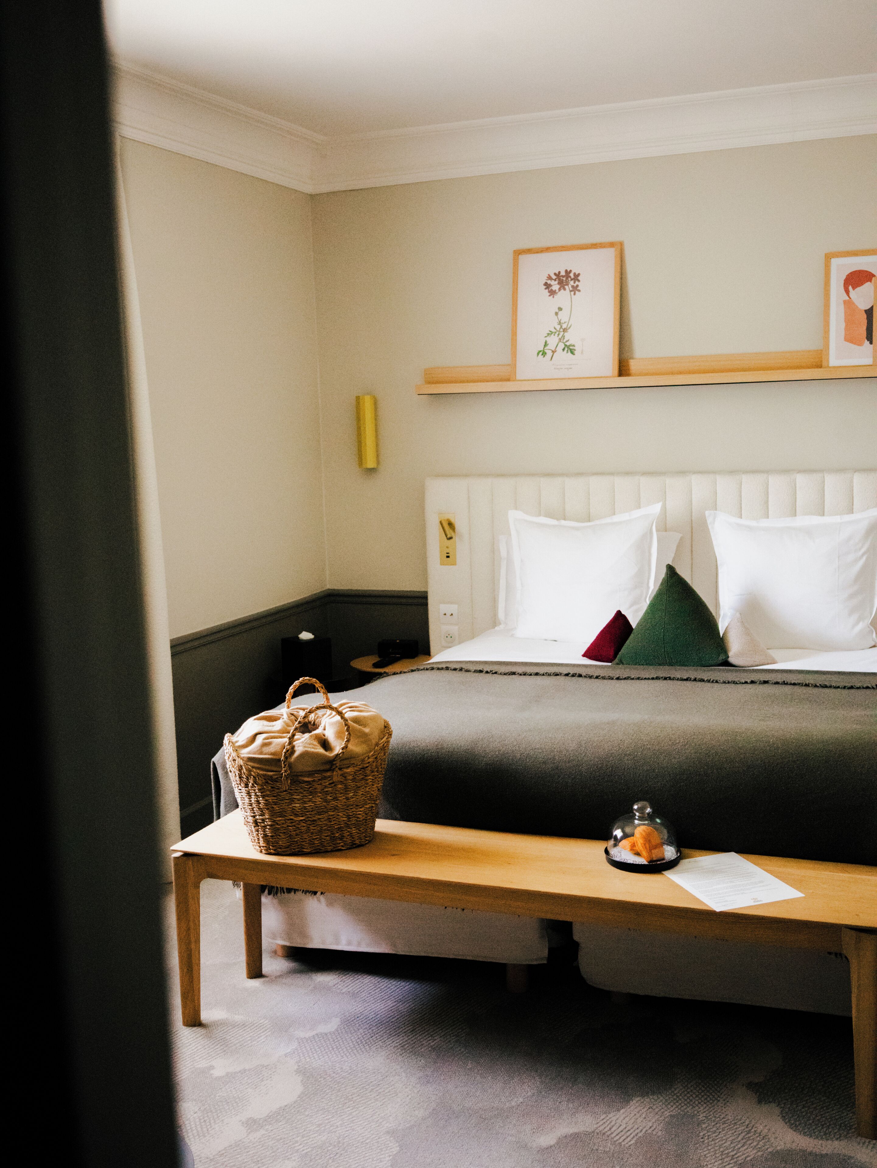 A neatly arranged hotel room with a bed, a bedside table, a bench, and a wall-mounted shelf with framed pictures.