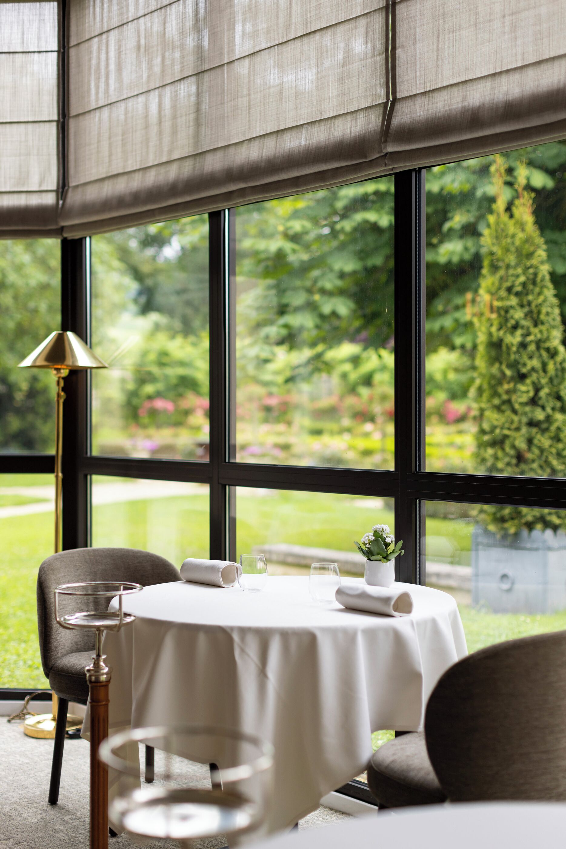Een eethoek met een tafel gedekt voor twee personen, een lamp en uitzicht op een tuin door grote ramen.