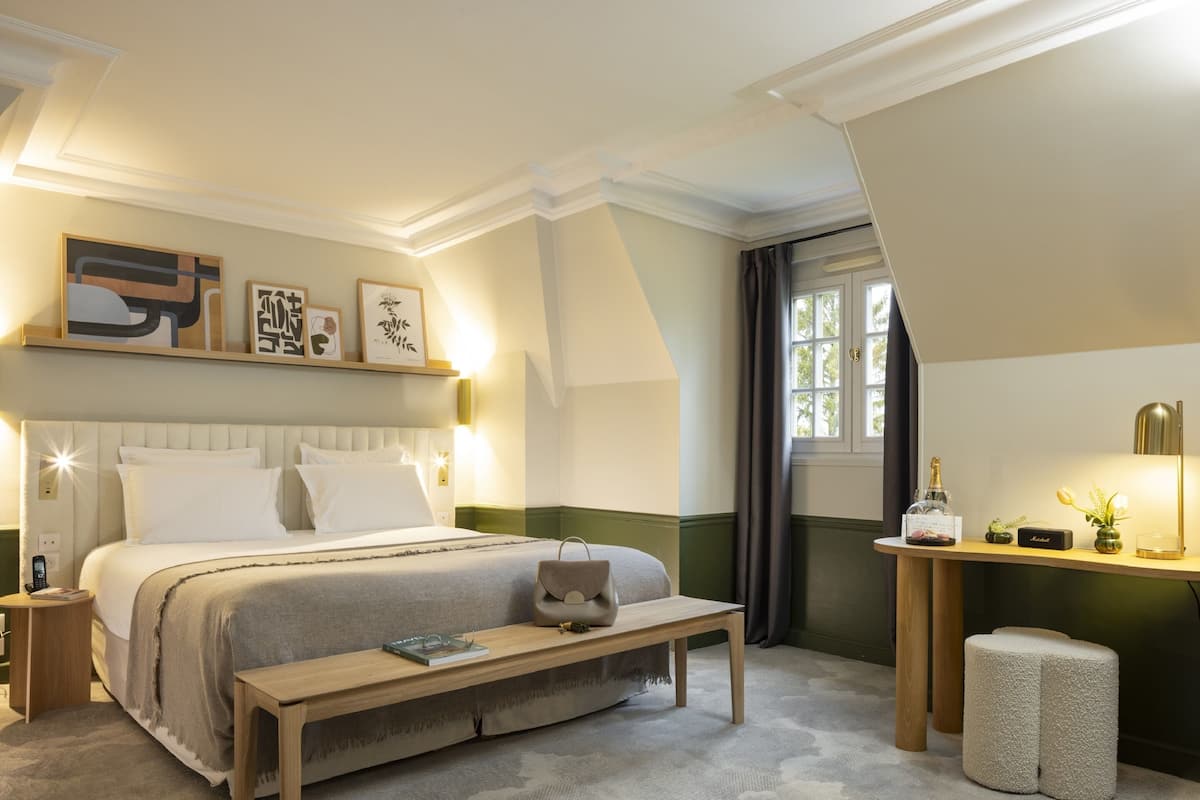 Een keurig opgemaakte slaapkamer met een bed, nachtkastjes, een bedlampje, een klein tafeltje met een plant en een raam met gordijnen.