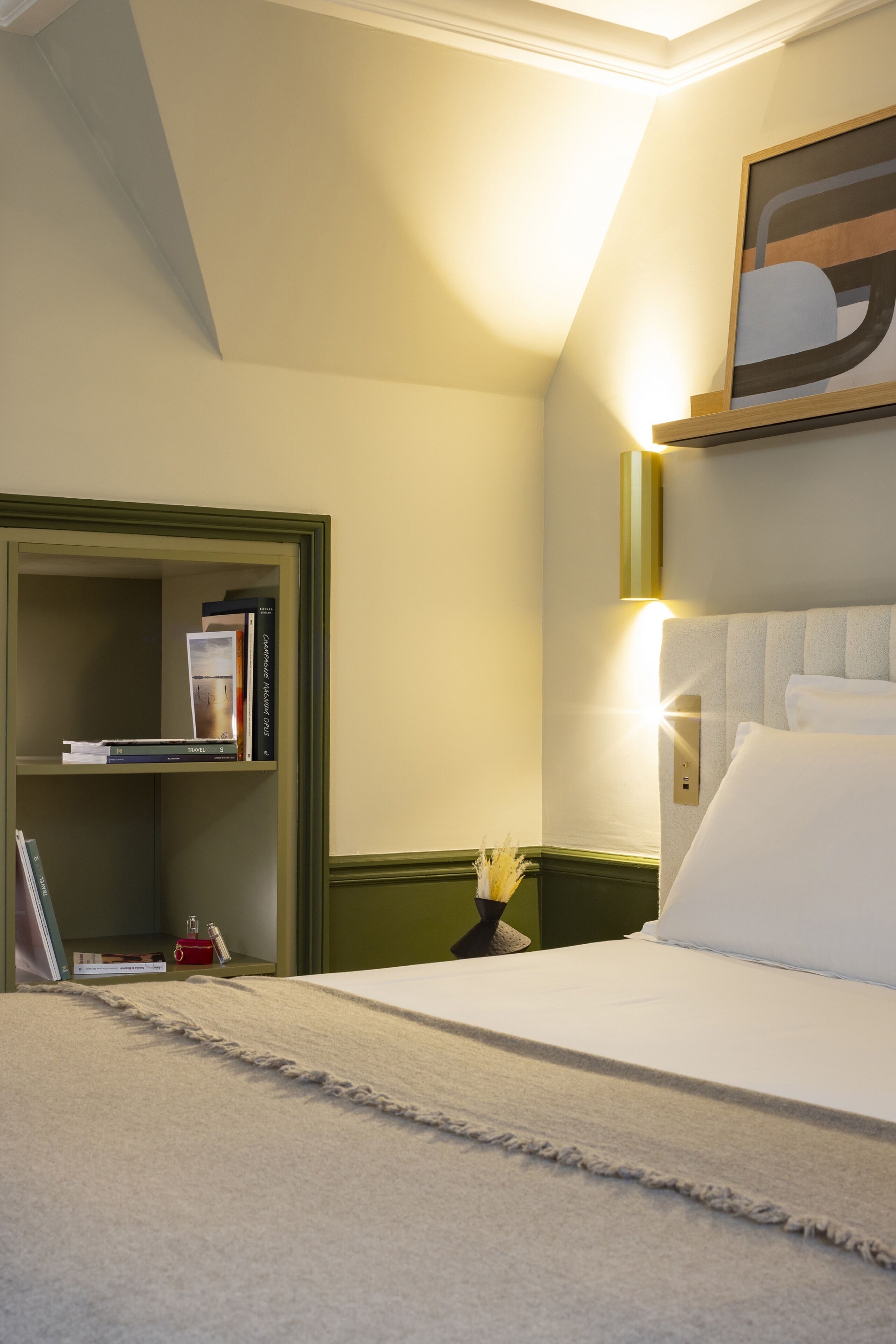A hotel room with a bed, a bookshelf, and a wall-mounted light fixture.