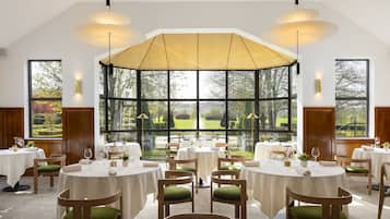 Un espace repas avec des tables rondes, des chaises rembourrées de couleur verte et une grande fenêtre donnant sur un jardin.