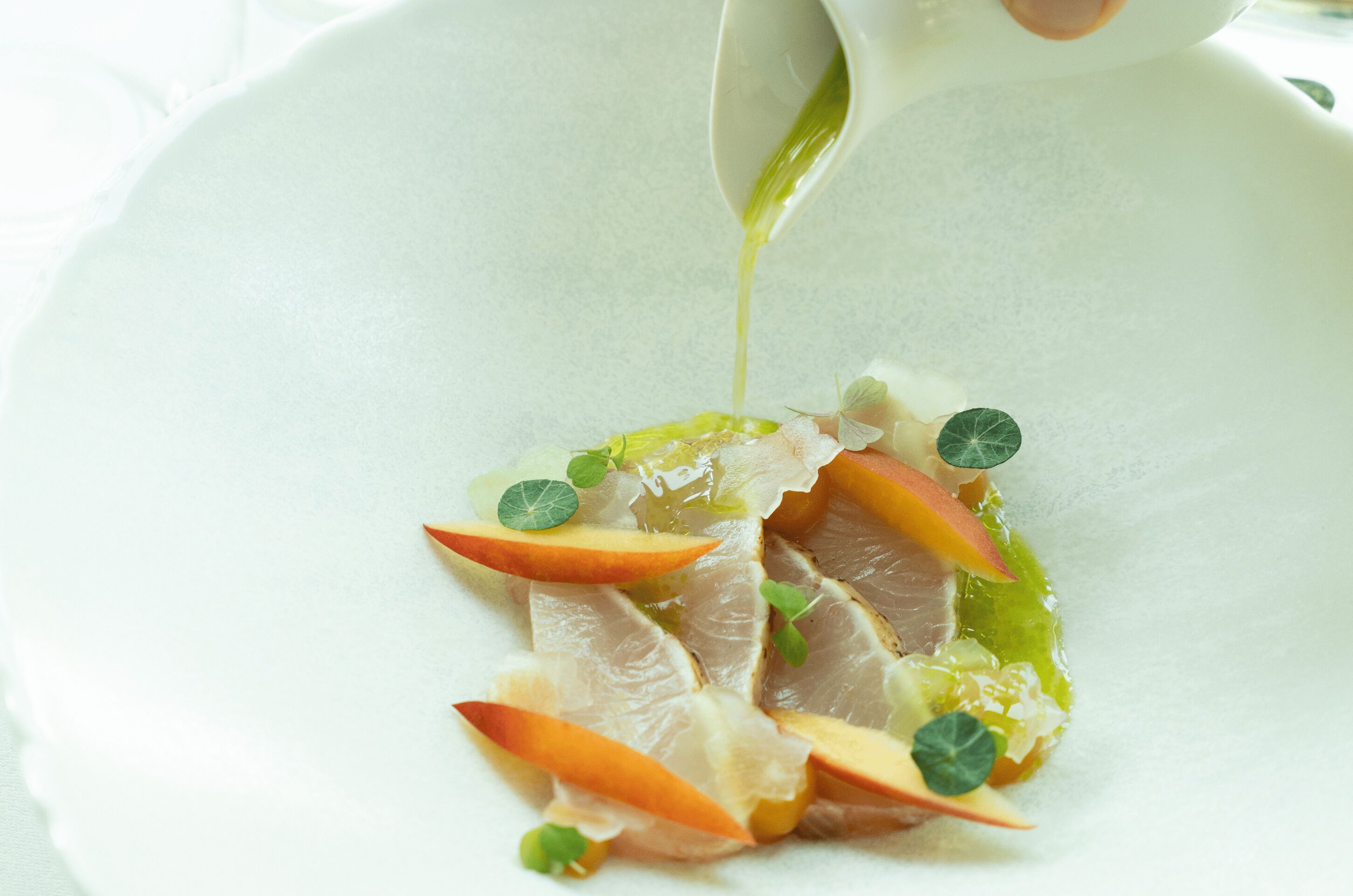 A white plate with thinly sliced meat, apple slices, and a green sauce being poured over the dish.