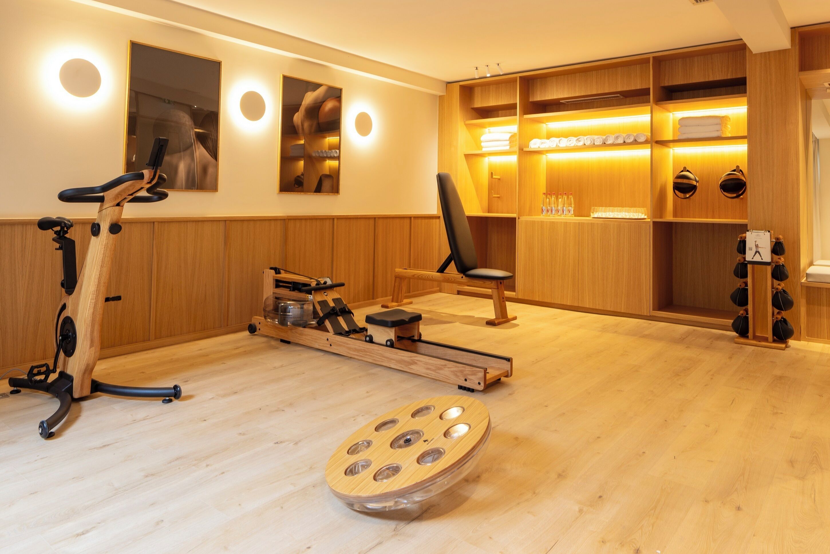 A room with a wooden floor, exercise equipment, a chair, and a shelf with towels and bottles.
