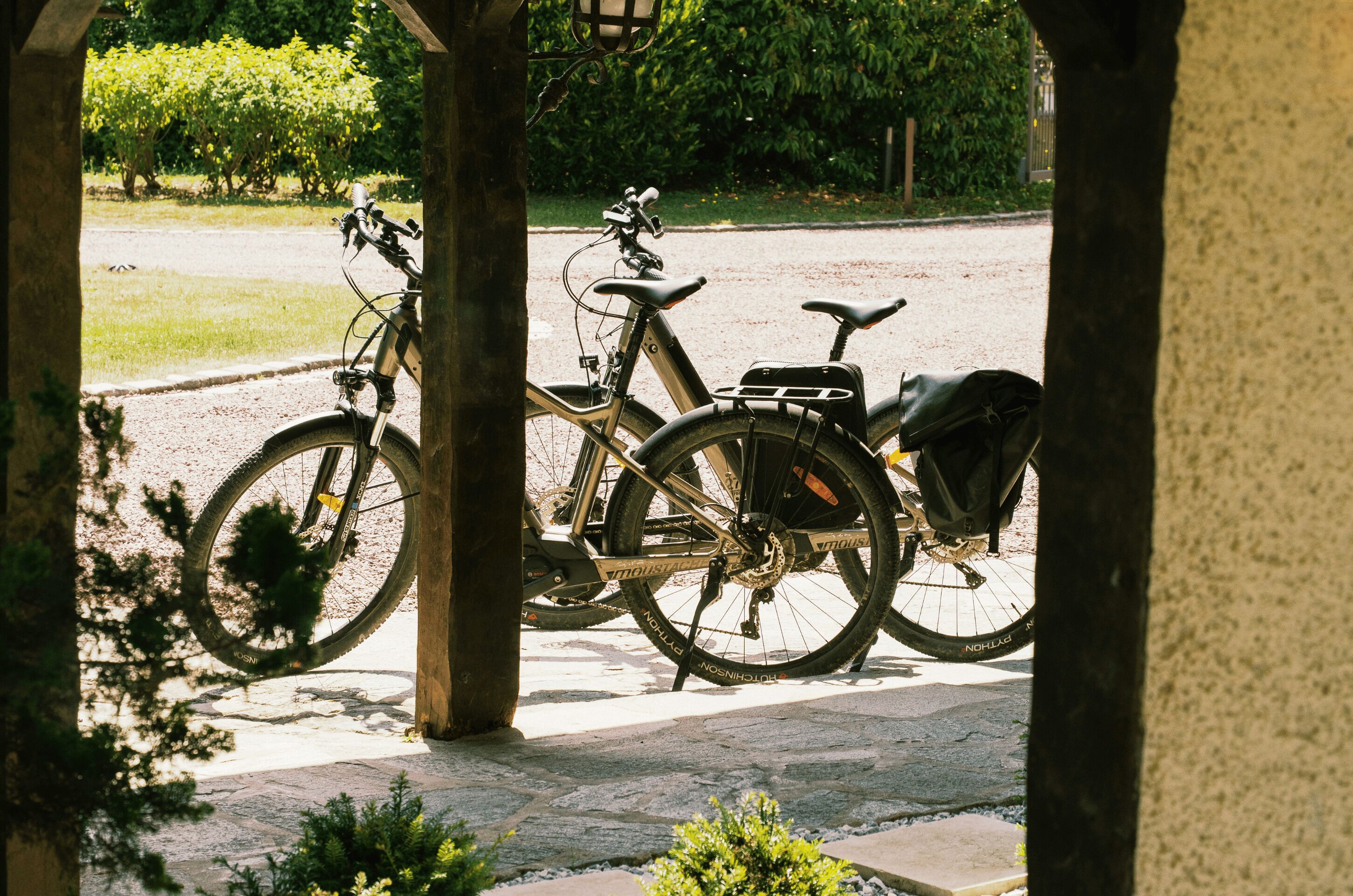 A bicycle parked under a covered area with a stone pavement and greenery in the background.