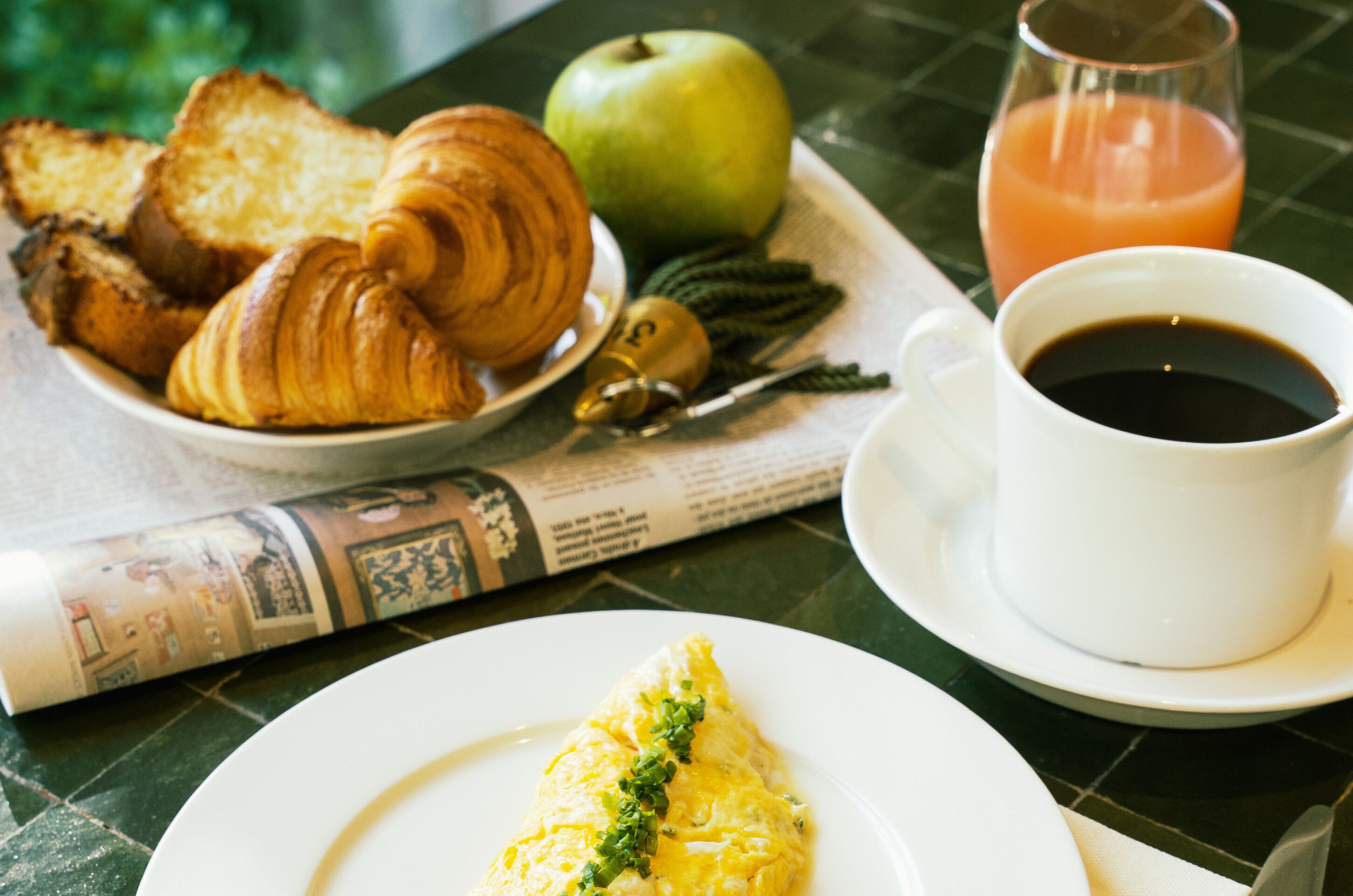 A breakfast setting with croissants, an apple, a glass of orange juice, a cup of coffee, and an omelette on a table.