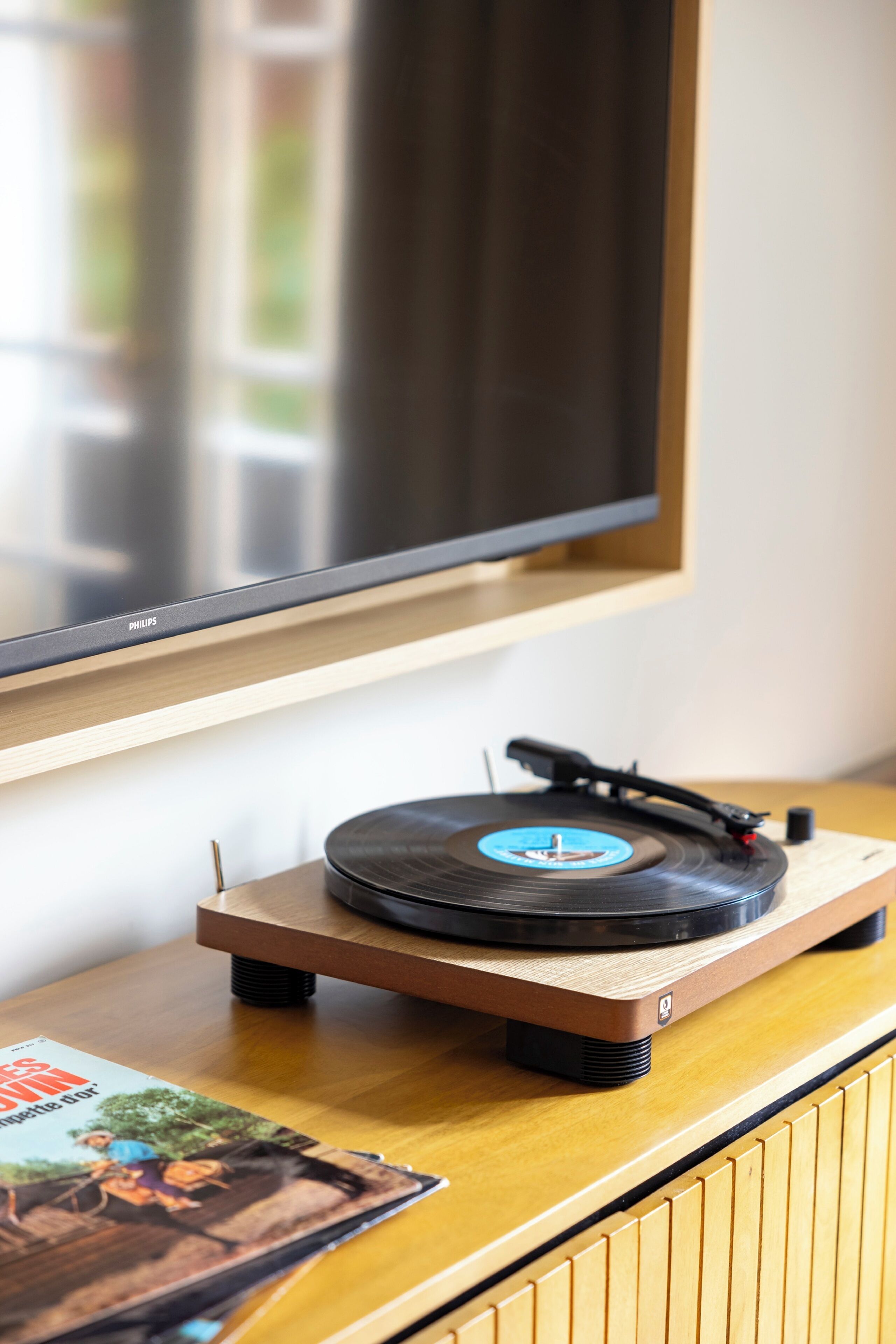 Tocadiscos sobre una mesa de madera con un disco de vinilo, un televisor montado en la pared y una revista sobre la mesa.