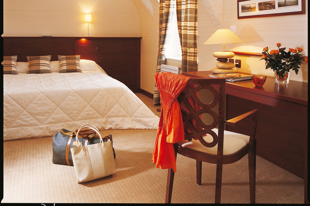 Ein Hotelzimmer mit einem Bett, einem Schreibtisch, einem Stuhl, einer Lampe, einer Vase mit Blumen und einer Handtasche auf dem Boden.