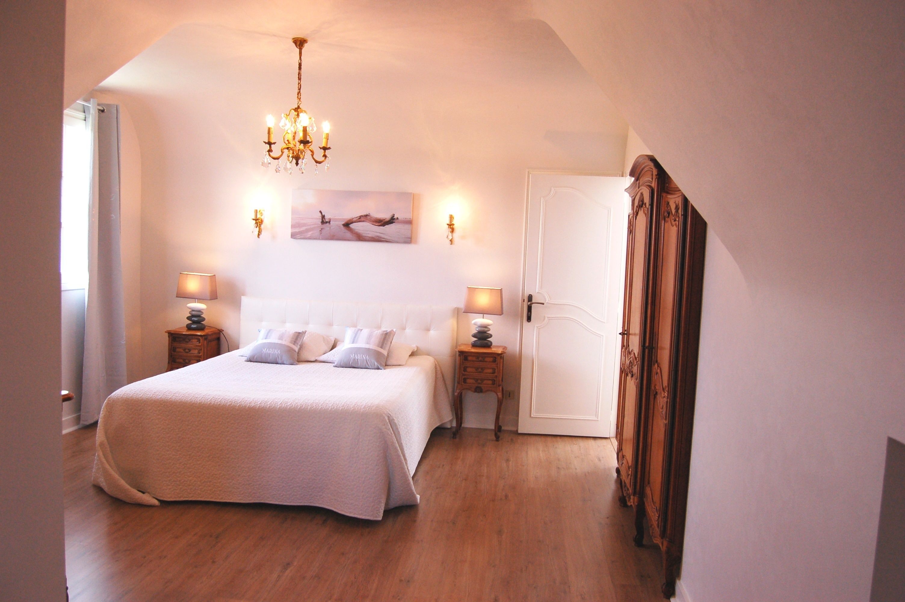 A bedroom with a large bed, two bedside tables with lamps, a chandelier, a door, and a painting on the wall.