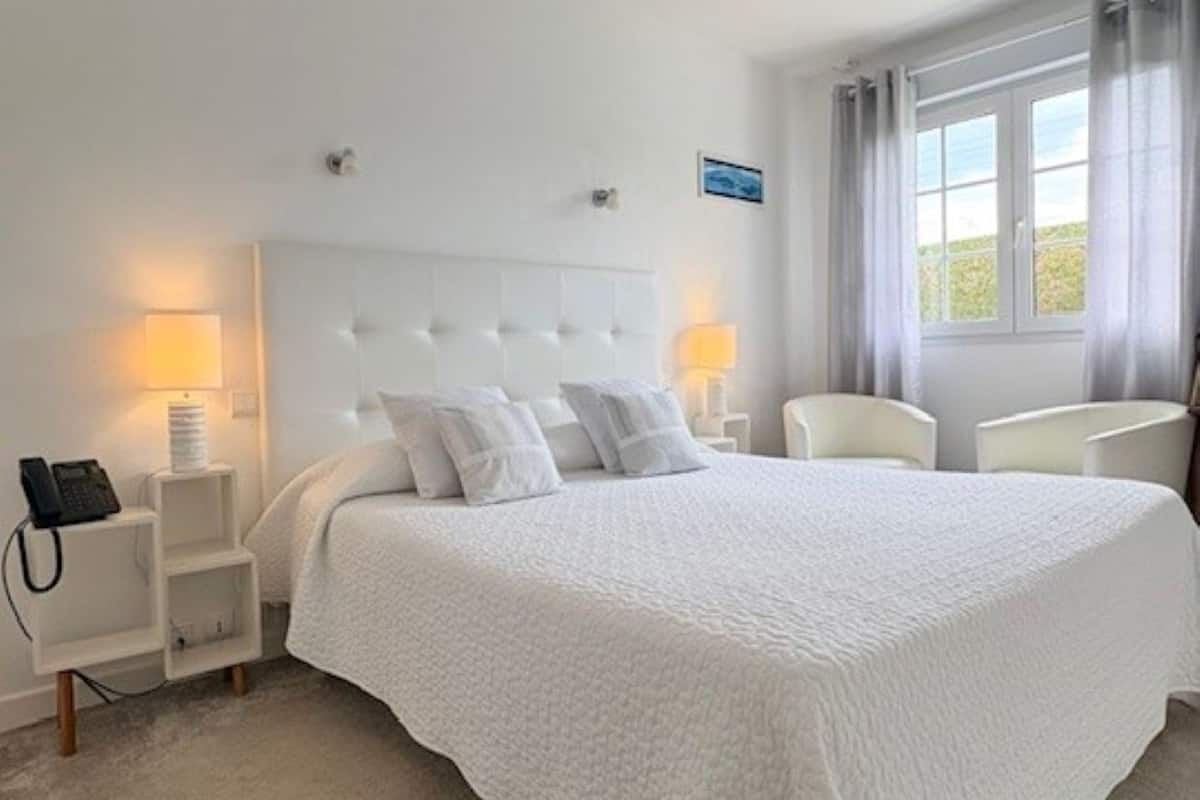 Ein Hotelzimmer mit einem großen Bett, Nachttischlampen, einem Sessel, einem Fenster mit Vorhängen und einem kleinen Tisch mit einem Telefon.