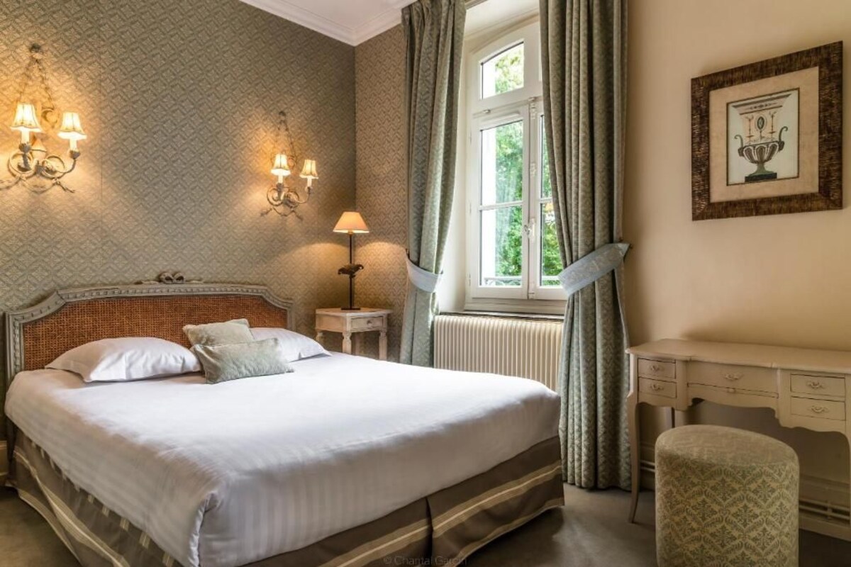 A neatly made bed with a headboard, a bedside table with a lamp, a window with curtains, and a framed picture on the wall.