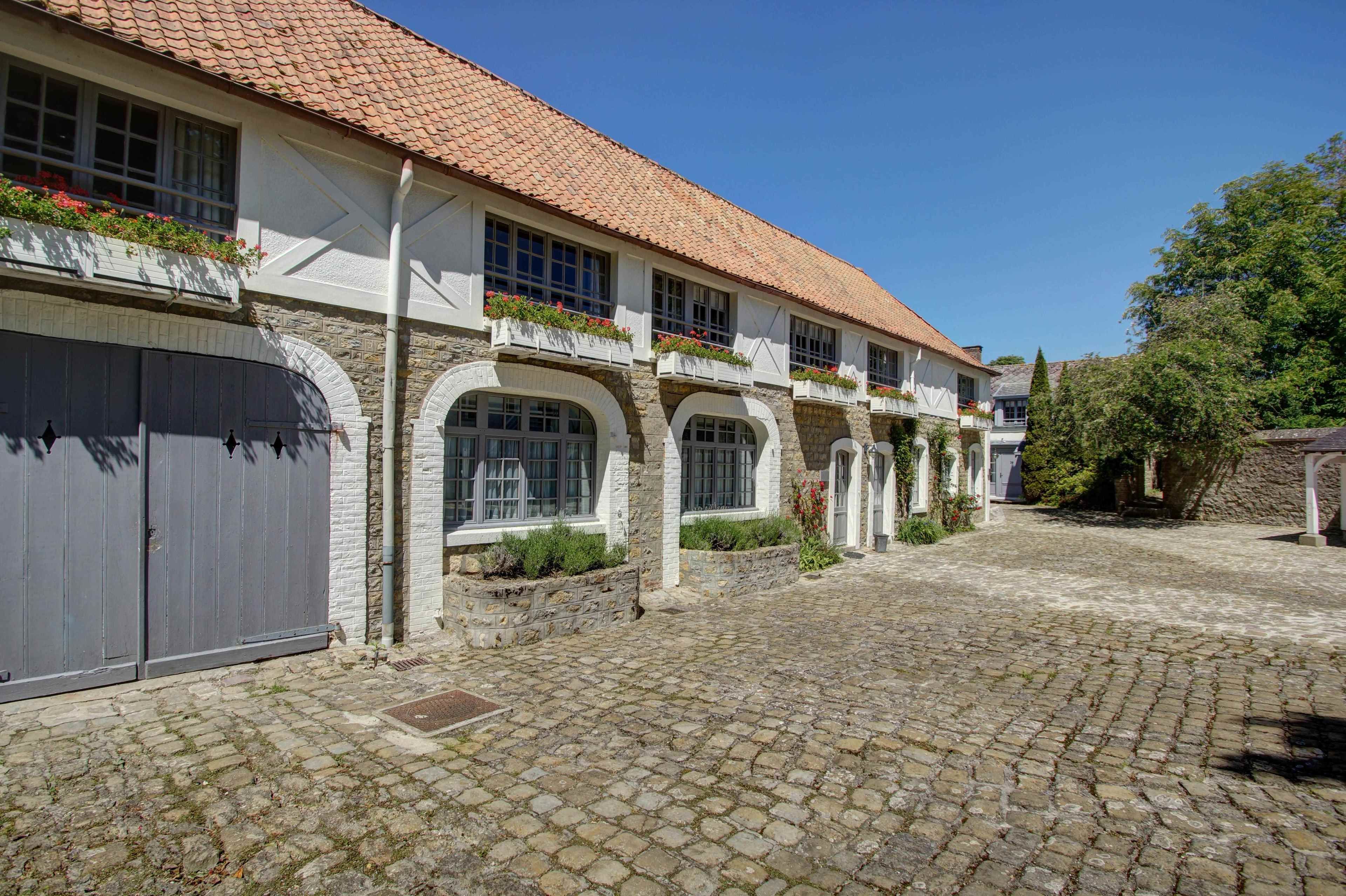 Une cour dallée de pierres, un bâtiment avec un toit en tuiles, des arches en pierre et une porte de garage grise.