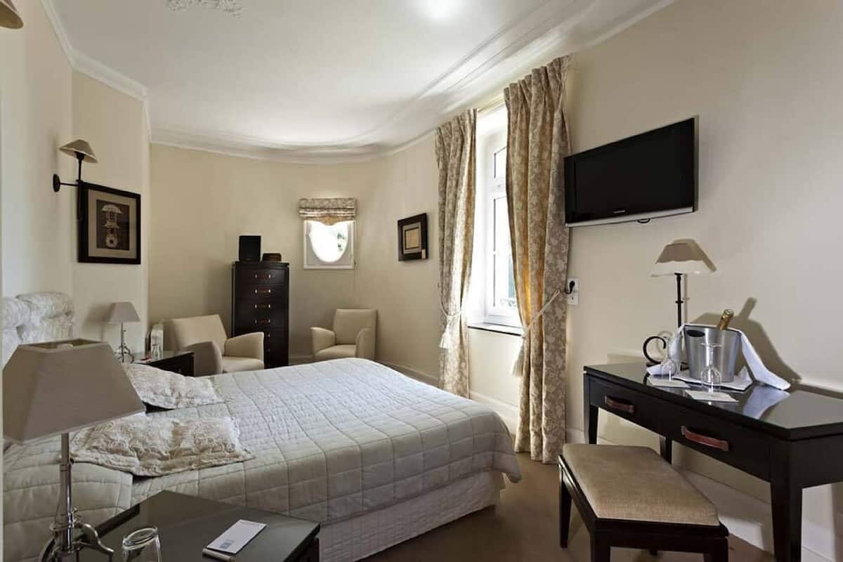 A hotel room with a bed, a desk with a chair, a television, a dresser, and a window with curtains.