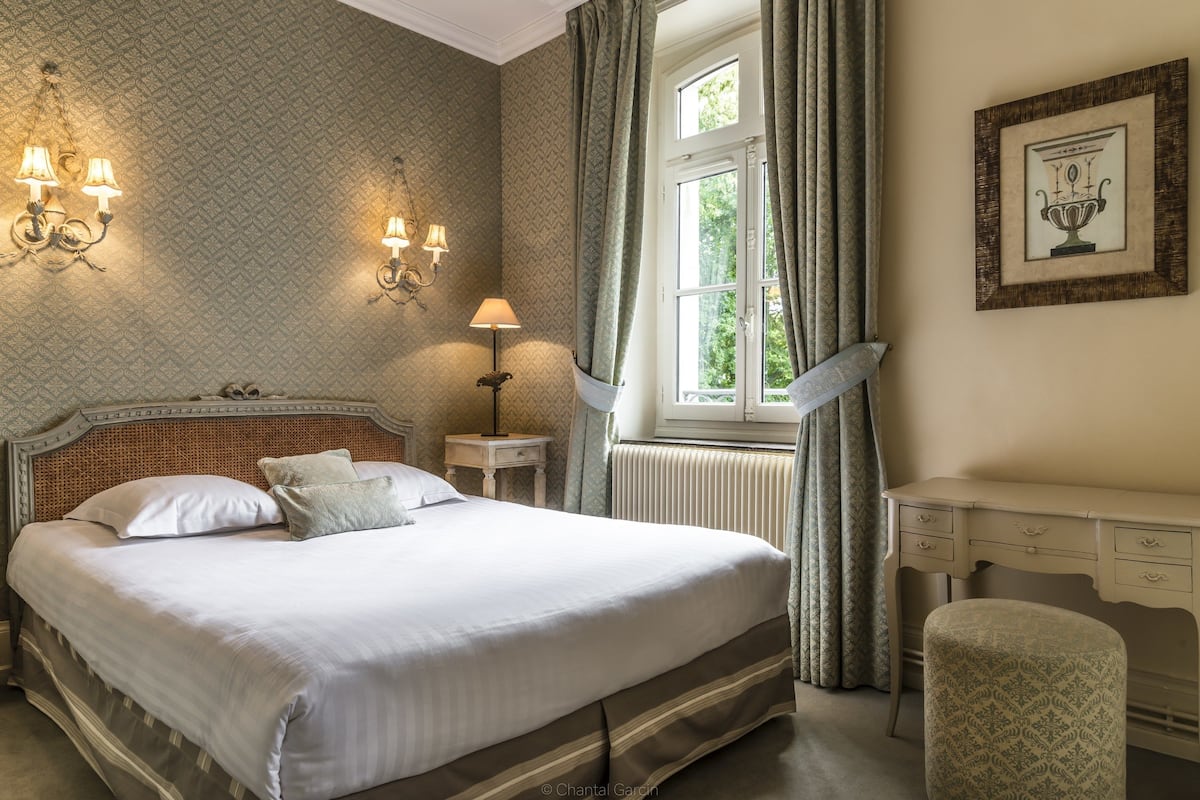 A neatly arranged bedroom with a large bed, a bedside table, a chair, and a window with curtains.