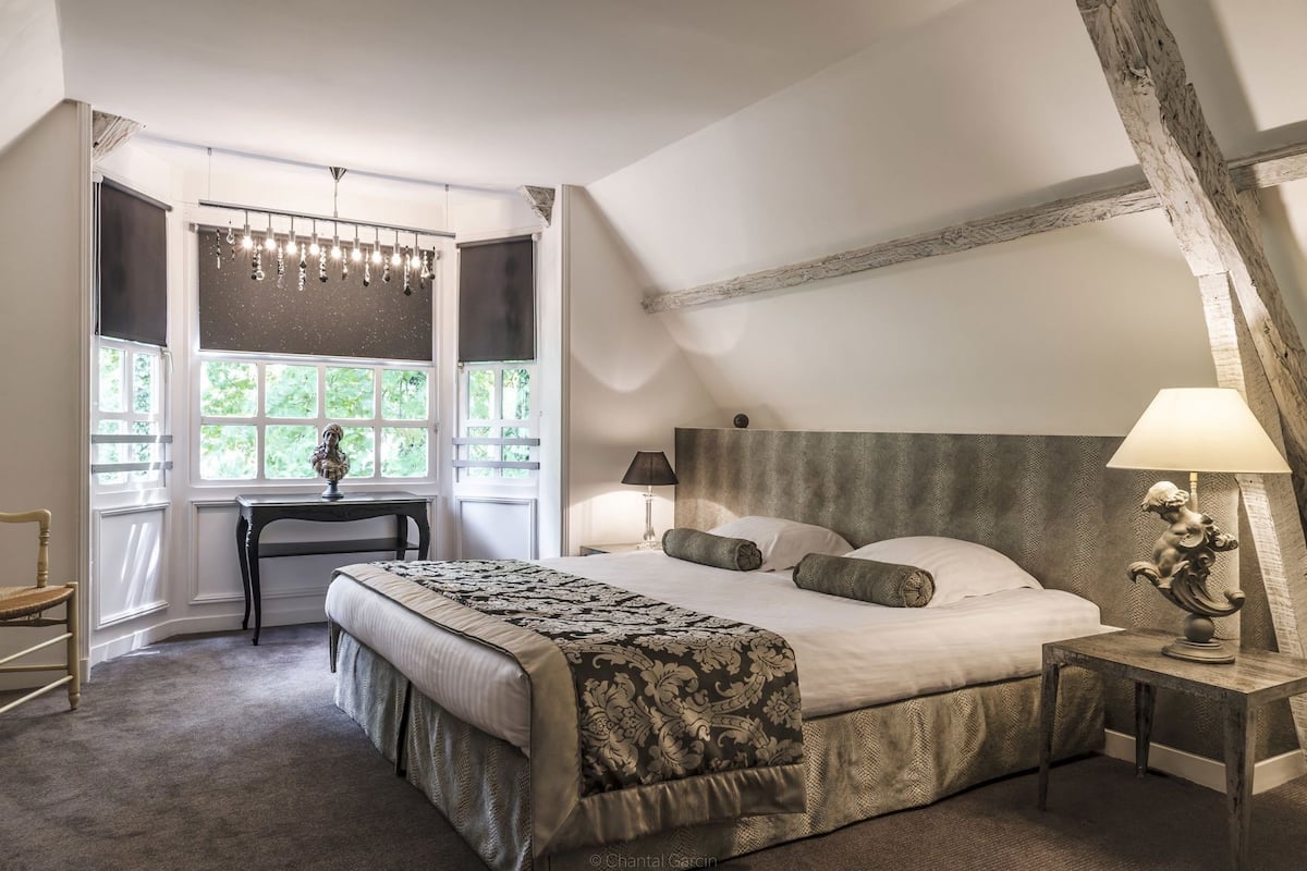 A bedroom with a large bed, a desk, a chair, and a window with a view of greenery.