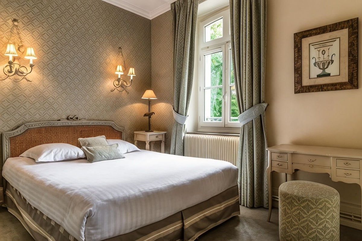 A hotel room with a large bed, bedside lamps, a window with curtains, a radiator, a desk, and a chair.