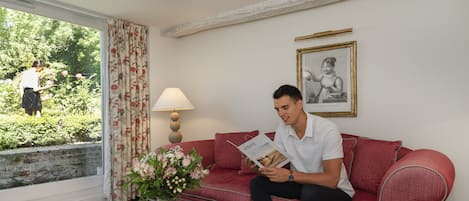 Um homem sentado em um sofá vermelho lendo uma revista, com um vaso de flores sobre uma mesa em frente a uma janela com cortinas.